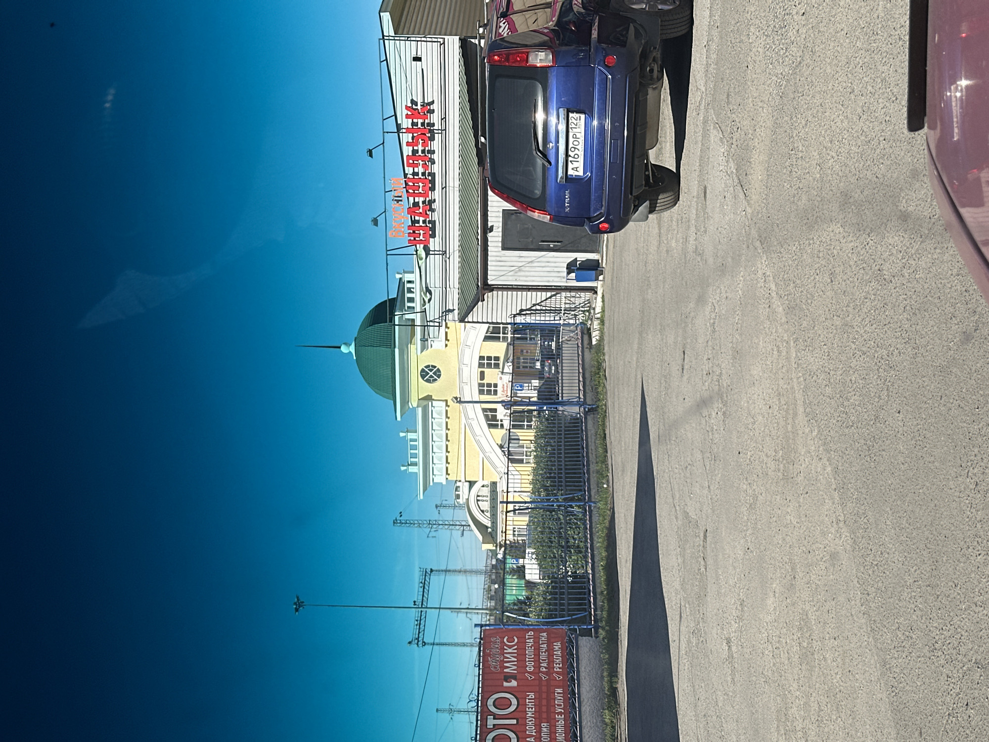 The image shows a street scene in Novoaltaysk, Russia, with a blue SUV parked in front of a building. The building appears to be a commercial establishment, possibly a restaurant or shop, with a sign in Russian that reads "Вкусный Шашлык" (Delicious Shashlik). There is also a red banner on the left side of the image with Russian text, possibly advertising services. In the background, a yellow building with a green dome is visible, along with power lines and other structures. The sky is clear and blue, suggesting it is a sunny day. The SUV has a license plate that reads "A 169OP 172". The ground is paved with asphalt.