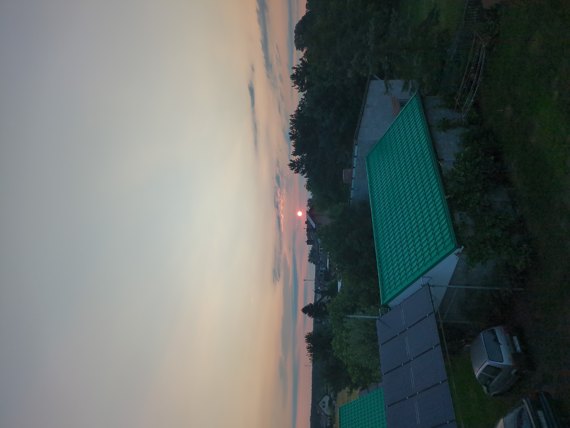 The image captures a sunset scene over the town of Rusiec, Poland. The sky is a gradient of soft pinks, oranges, and pale blues, with thin, wispy clouds scattered across it. The sun, a small, reddish-orange orb, is visible near the horizon, partially obscured by clouds. In the foreground, several buildings with vibrant green roofs are prominent. One building appears to be a garage or shed, with solar panels installed on its roof. Adjacent to it is another structure with a similar green roof. A silver car is parked beneath the solar panels. The surrounding area is densely populated with trees, forming a thick green canopy that extends towards the horizon. In the distance, houses with various roof types are visible amidst the trees, indicating a residential area. The overall impression is one of a tranquil evening in a semi-rural setting.