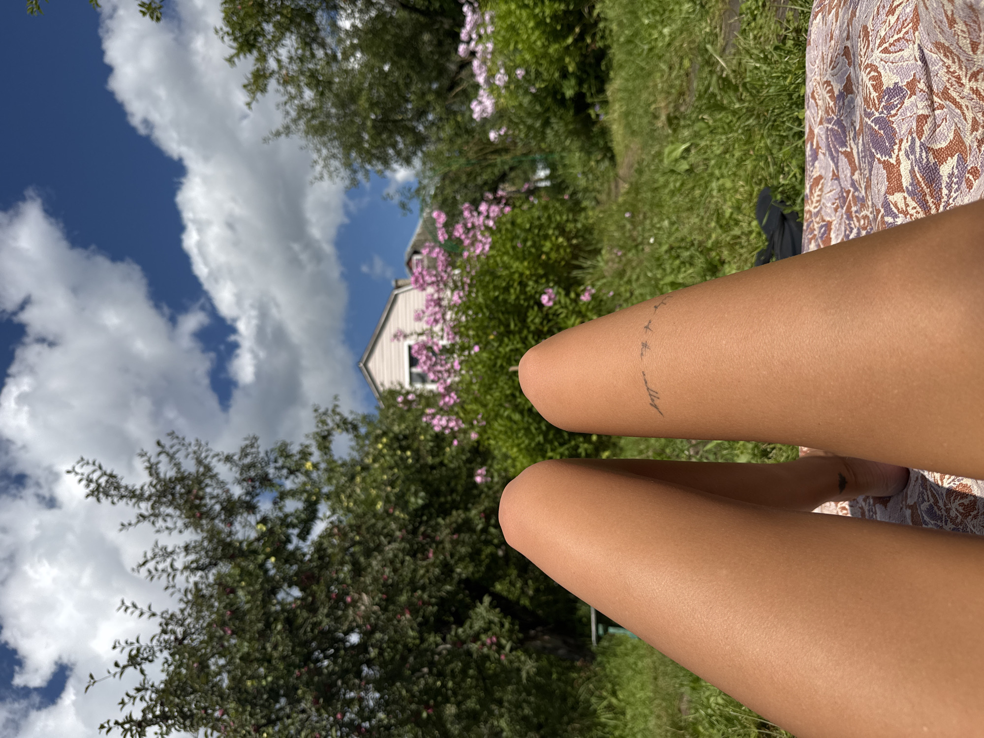 The image shows a first-person perspective of someone lying on a patterned blanket in a grassy yard. The foreground is dominated by the person's tanned legs, one of which has a faint tattoo of birds in flight. In the background, a lush garden is visible with a variety of green foliage and blooming pink flowers. Beyond the garden, a light-colored house with a pink roof is partially visible, set against a bright blue sky with fluffy white clouds. The environment suggests a warm, sunny day in a rural or suburban setting, possibly a backyard in Красная Пресня Снт, Russia, given the context.