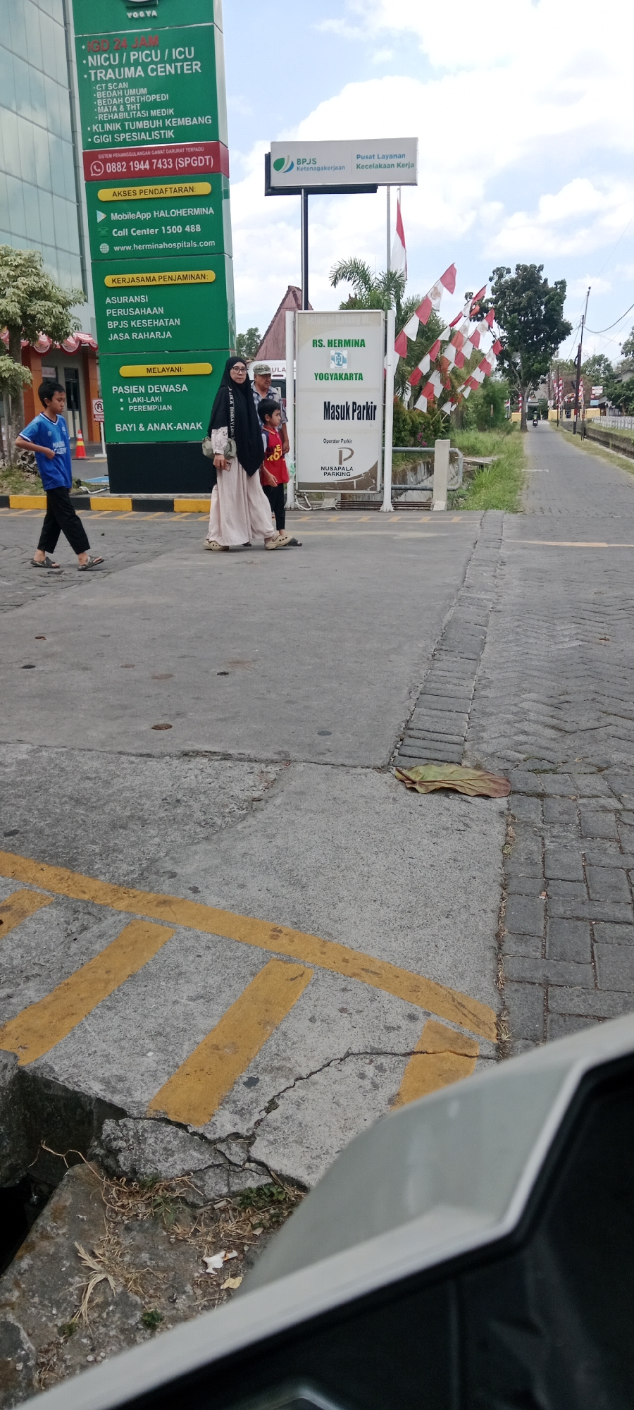 In Maguwoharjo, Indonesia, a street scene unfolds under a bright sky with scattered clouds. A large green and white sign for Hermina Hospitals Yogyakarta stands prominently on the left. To its right, a smaller sign indicates "RS. HERMINA YOGYAKARTA" with "Masuk Parkir" (Enter Parking) below. Red and white flags are strung across the scene.
On the pavement in front of the hospital signs, three people are walking: a young boy in a blue shirt and dark pants, followed by a woman in a cream-colored abaya and black hijab, and a young boy in a red shirt. A blue and white sign with "BPJS Ketenagakerjaan" (BPJS Employment) is visible to the right of the hospital sign. The foreground shows cracked concrete and yellow road markings. The road stretches into the distance, lined with greenery and buildings. The scene is captured from a low angle, possibly from a vehicle.