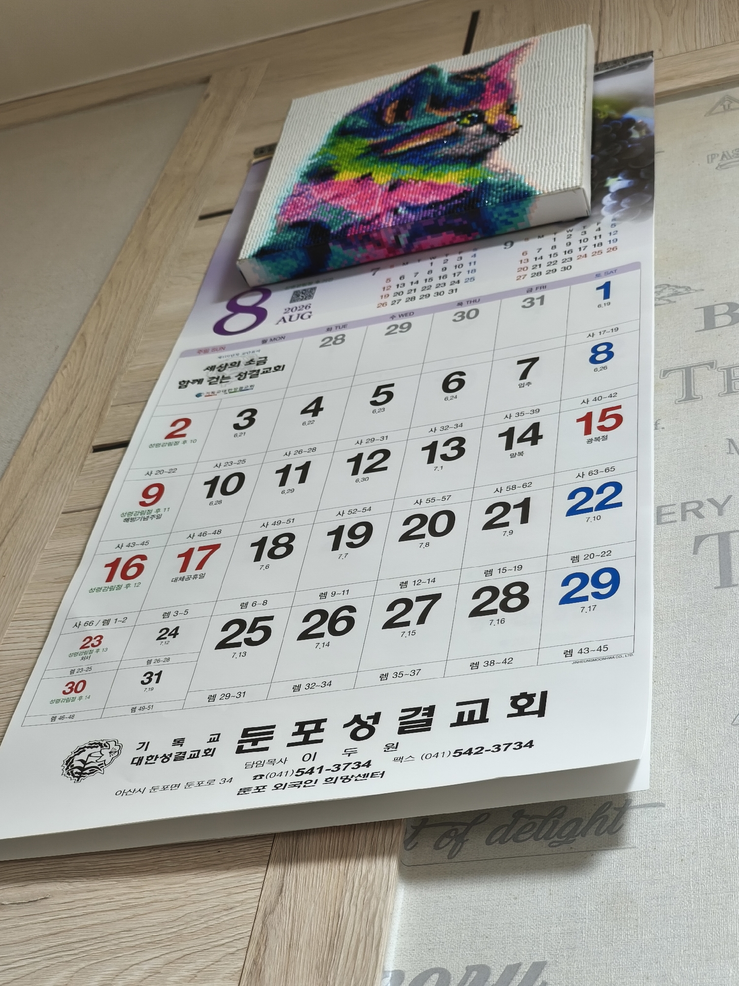 The image shows a wall calendar from August 2026, hanging on a light-colored wooden wall. The calendar is for the Tepo Sungkyul Church in Asan-si, South Korea. The calendar features a diamond painting of a cat at the top. Each day of the month is listed with a corresponding number and sometimes additional text. The bottom portion of the calendar displays the church's name, address (located in Dunan-myeon, Asan-si), phone number, and fax number. The lighting suggests an indoor environment, possibly a home or office.