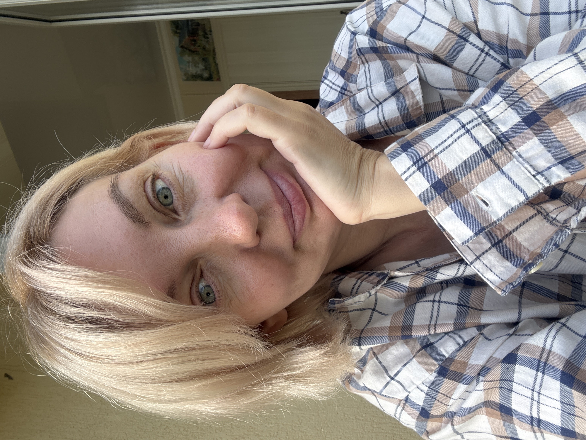 A woman with blonde hair and green eyes is taking a selfie outdoors. She is wearing a plaid shirt and resting her chin on her hand. She is smiling slightly. The background is blurry but appears to be an outdoor setting with a wall and a window. The image quality is good, and the lighting is natural. There is no indication of the location in the city of Sochi, Russia.