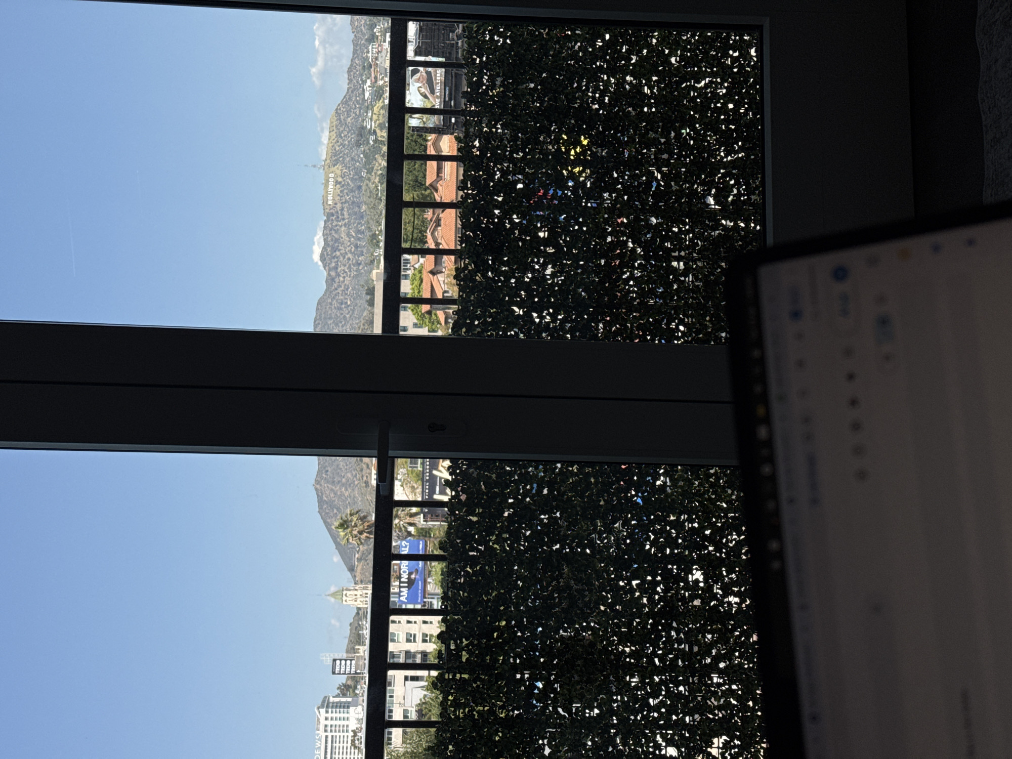 A view from an open window of a building in Los Angeles, United States, shows a cityscape and mountains on a clear day. The Hollywood sign is visible on the hillside in the background. A faux hedge covers part of the window. A computer screen is in the foreground.
