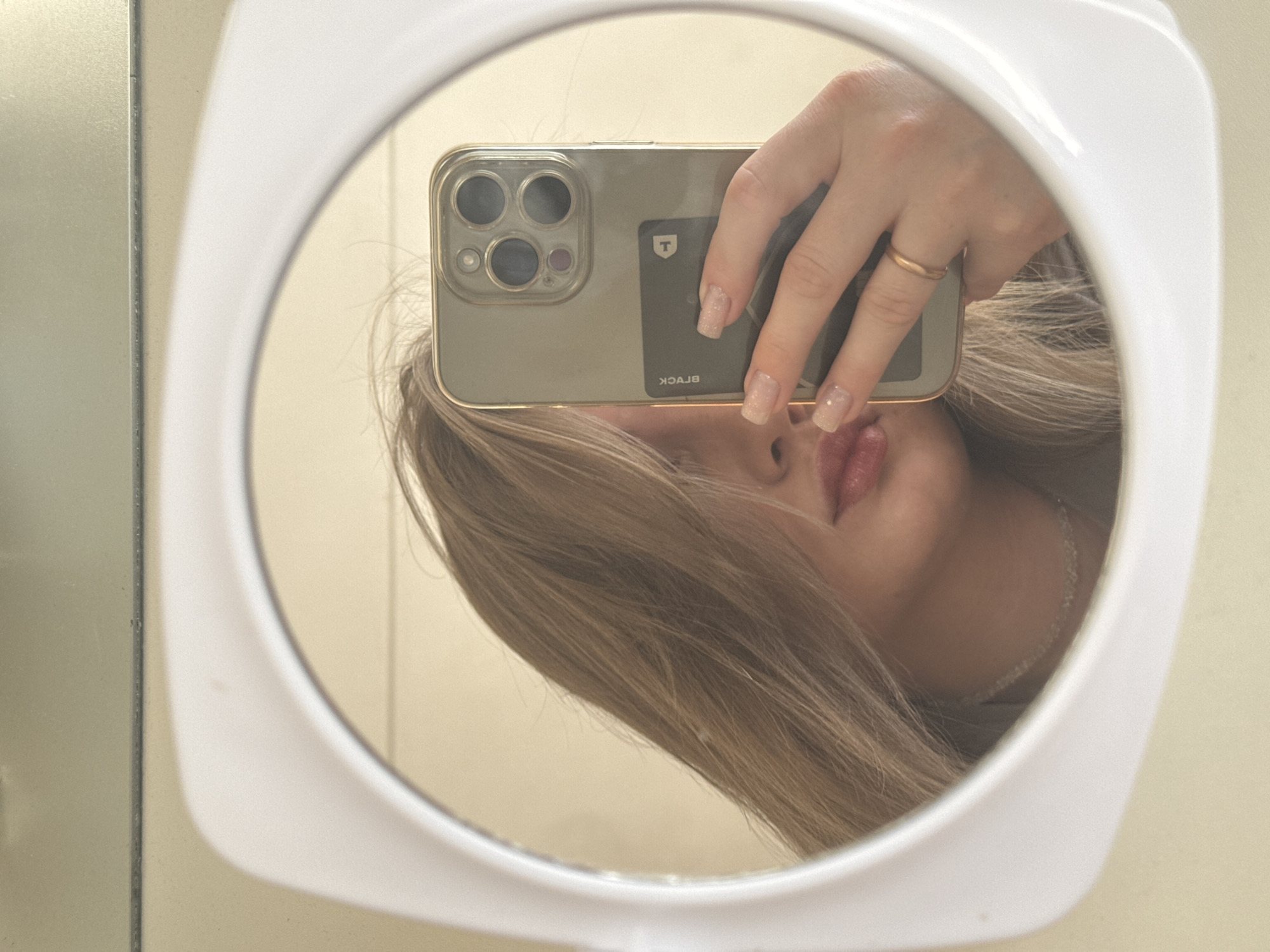 A woman with blonde hair is taking a selfie in a white-framed round mirror. She is holding a gold iPhone with multiple lenses and a black card attached to the back. She is wearing a gold ring on her finger. The image appears to be taken indoors, against a light-colored wall. The location of Chaikovsky, Russia, is not definitively supported by the visual information provided.