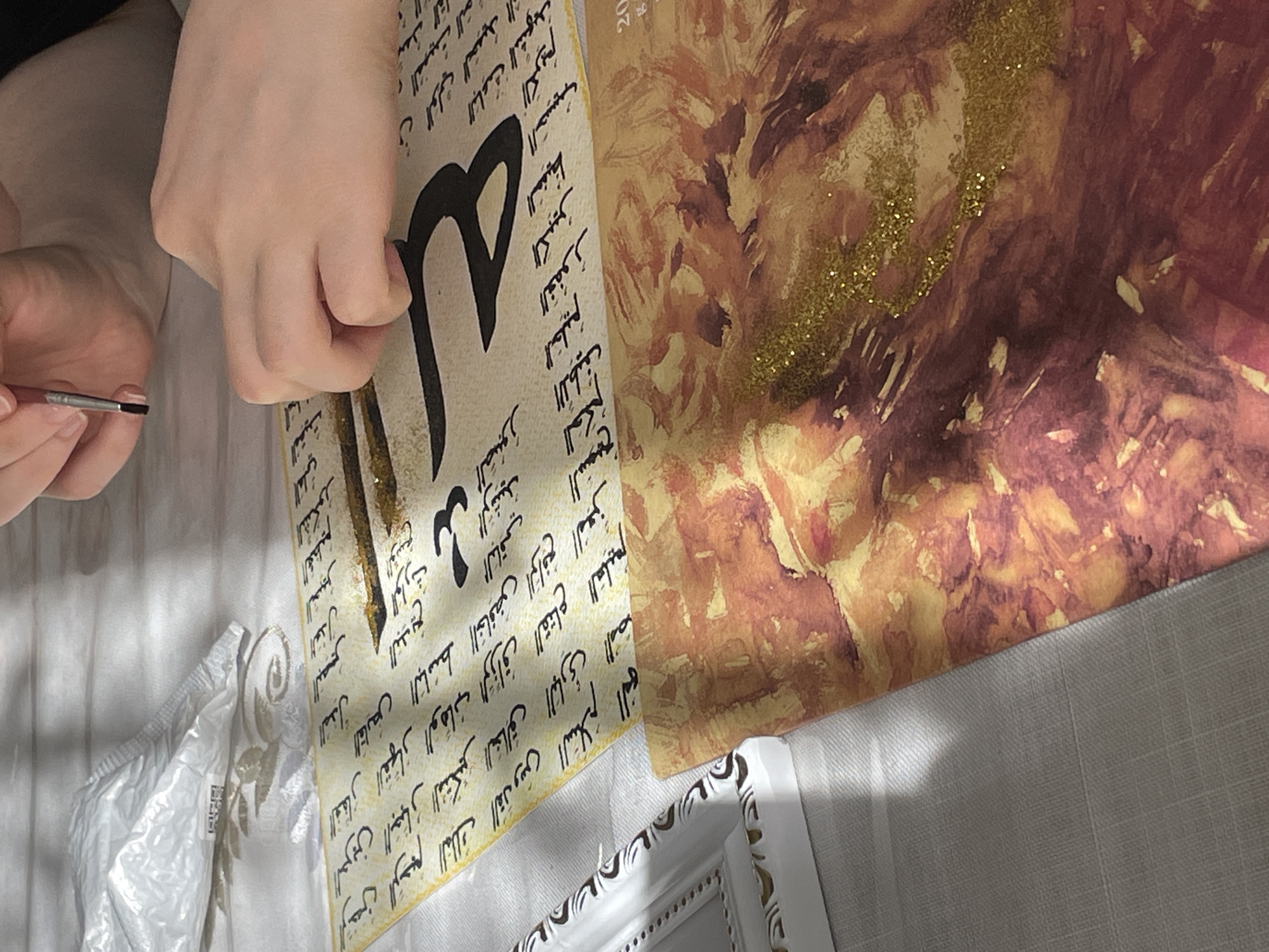 The image shows a close-up of hands engaged in artistic creation on a white tablecloth. A person is holding a fine paintbrush with their right hand, while their left hand is placed on a paper adorned with black Arabic calligraphy. The calligraphy is written in several rows, with some larger, bold strokes interspersed, possibly forming a decorative element or a prominent word. Gold glitter is scattered on the paper, catching the light. Below this paper, another sheet with an abstract watercolor painting in warm tones of orange and red is visible, also with hints of gold glitter. A white decorative frame with a brown pattern is positioned to the left of the main subject. In the upper left corner, a white padded envelope is present. The overall scene suggests an indoor setting, likely a craft or art studio, with soft lighting casting shadows. There is no discernible information or visual cue in the image that places the scene in Tashkent, Uzbekistan.