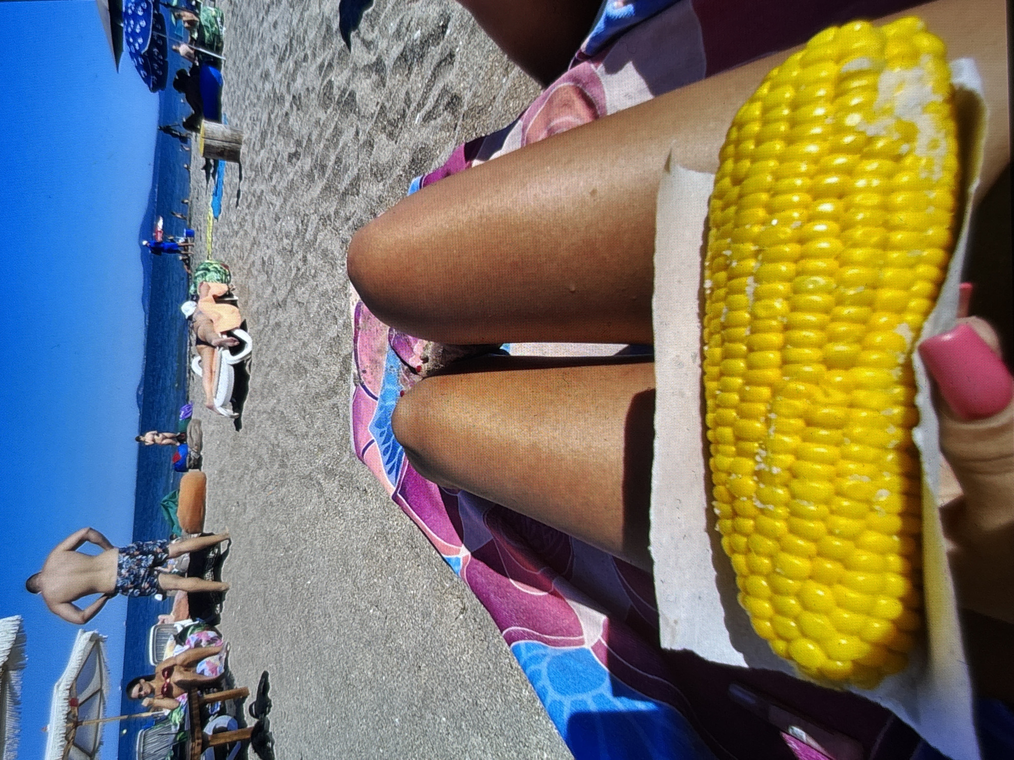 The image shows a first-person perspective of someone sitting on a beach towel, with their legs prominently in the foreground. A cob of corn, sprinkled with salt, rests on a white paper in front of their legs. The beach appears to be sandy, with other beachgoers in the background, some lounging on chairs and under umbrellas. The sea and a clear blue sky are visible beyond the beach. The location is identified as Burgas, Bulgaria.