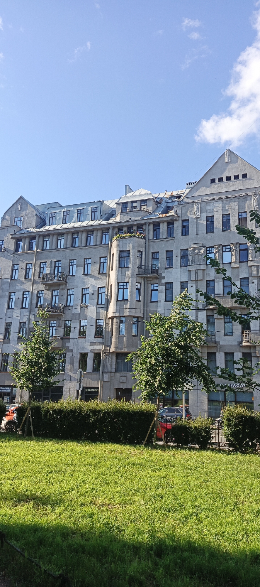 The image captures an exterior view of a multi-story building in Saint Petersburg, Russia. The building, with its distinctive gray facade and numerous windows, appears to be designed in an older architectural style, possibly Art Nouveau or Art Deco. Several trees are present in the foreground and mid-ground, some with supporting stakes. A neatly trimmed hedge runs along the front of the building, and a patch of green grass is visible in the lower portion of the frame. Cars are partially visible on the street behind the hedge. The sky above is a clear blue with scattered white clouds. The overall scene suggests a bright, sunny day in an urban setting.