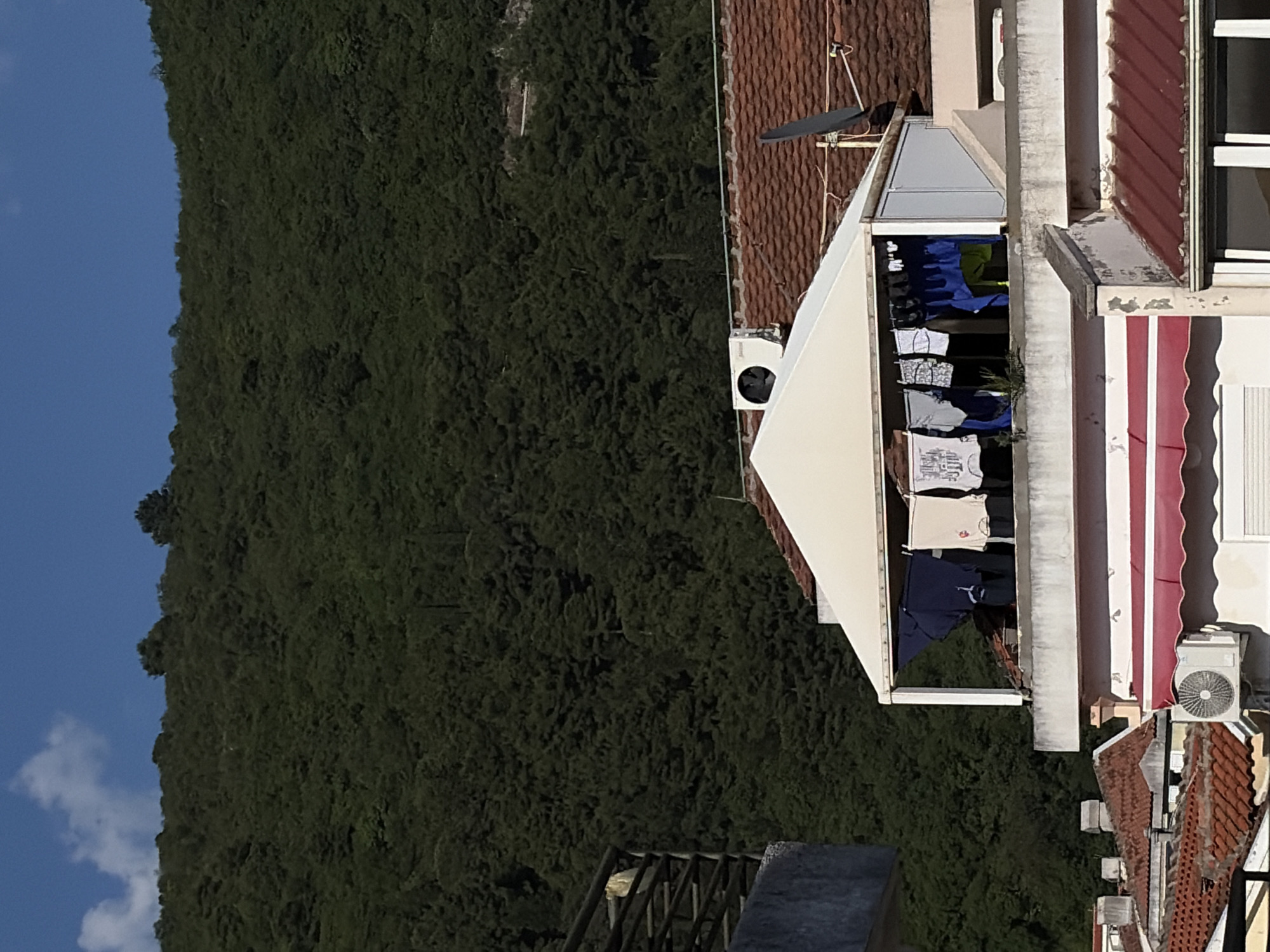 The image shows a view from above in Budva, Montenegro. A hillside covered in dark green vegetation fills the left side of the frame, contrasting with the clear blue sky at the top left. The right side features a building with red tile roofing, white walls, and a red and white striped awning. Laundry hangs within a covered balcony-like structure attached to the building. An air conditioning unit is visible on the building exterior. There are also other buildings with similar red tile roofs in the background, suggesting a dense urban area.