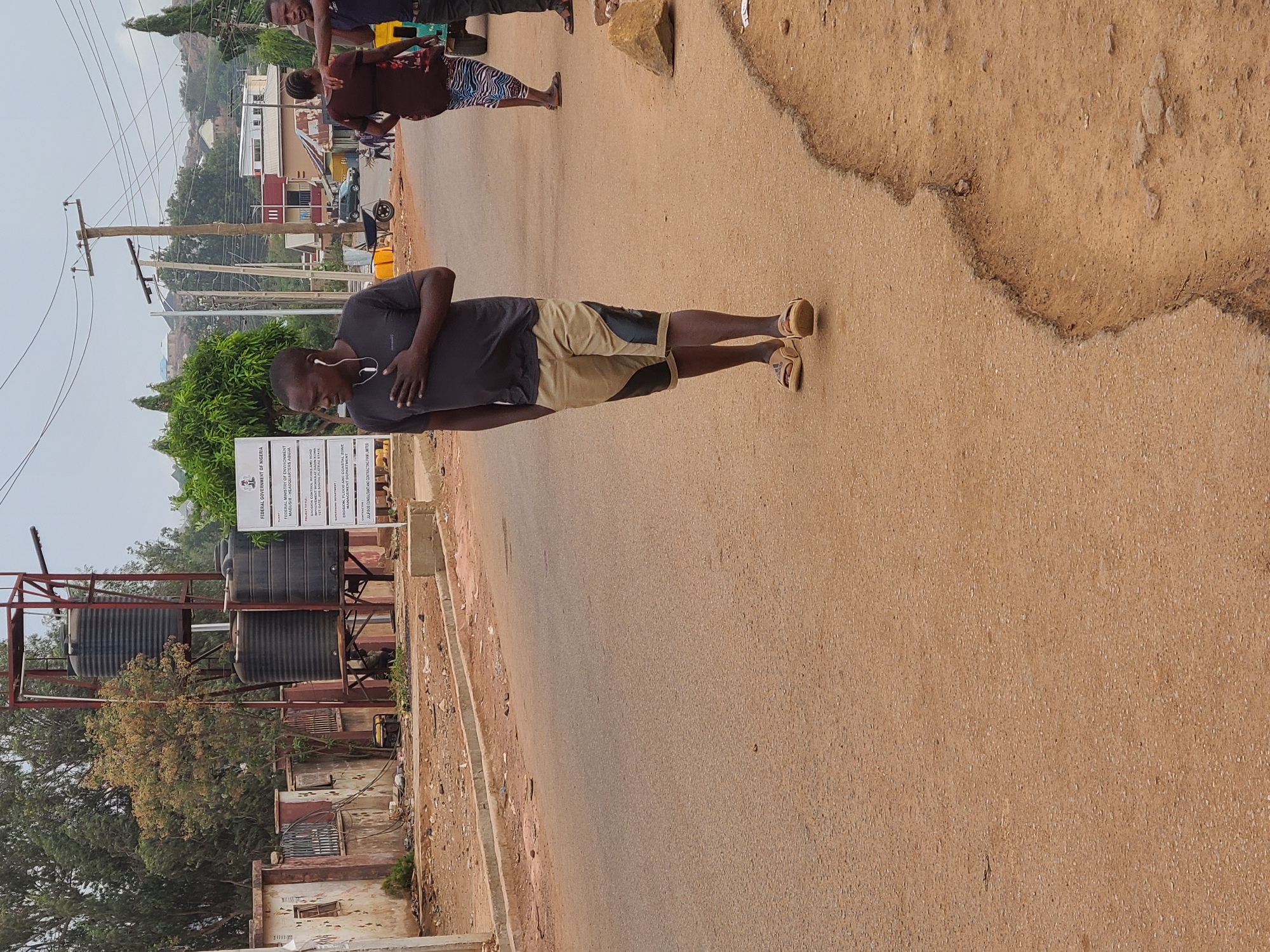 A man is standing on a street next to a building with large water tanks in Jos, Nigeria. The street is paved and shows signs of wear and tear. A sign is visible beside the tanks, indicating a "FEDERAL GOVERNMENT OF NIGERIA" project, specifically related to the "MINISTRY OF ENVIRONMENT" at "FEDEBUSIN HEADQUARTERS ABUJA". The sign further details the project as "EROSION CONTROL WORKS AND ROAD" at the "1ST GATE JOS SOUTH PLATEAU STATE". The man is wearing a dark shirt and khaki shorts with headphones, and he is looking away from the camera with his hand on his chest. In the background, other people are standing on the road. Buildings and vegetation are visible further in the distance, suggesting an urban or semi-urban environment. There are power lines in the background.
