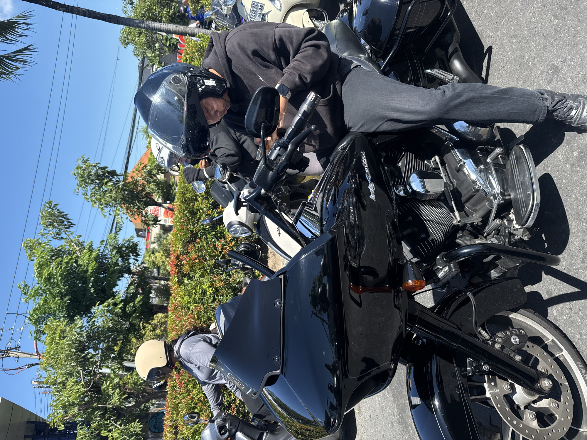 The image shows a man in a black hooded sweatshirt and full-face helmet sitting on a black motorcycle in Denpasar, Indonesia. He appears to be adjusting the controls or looking at the dashboard of the motorcycle. The motorcycle is parked on a paved road alongside other motorcycles and scooters. In the background are buildings, trees, and other vegetation typical of a city street in Denpasar. The weather appears to be sunny and clear.