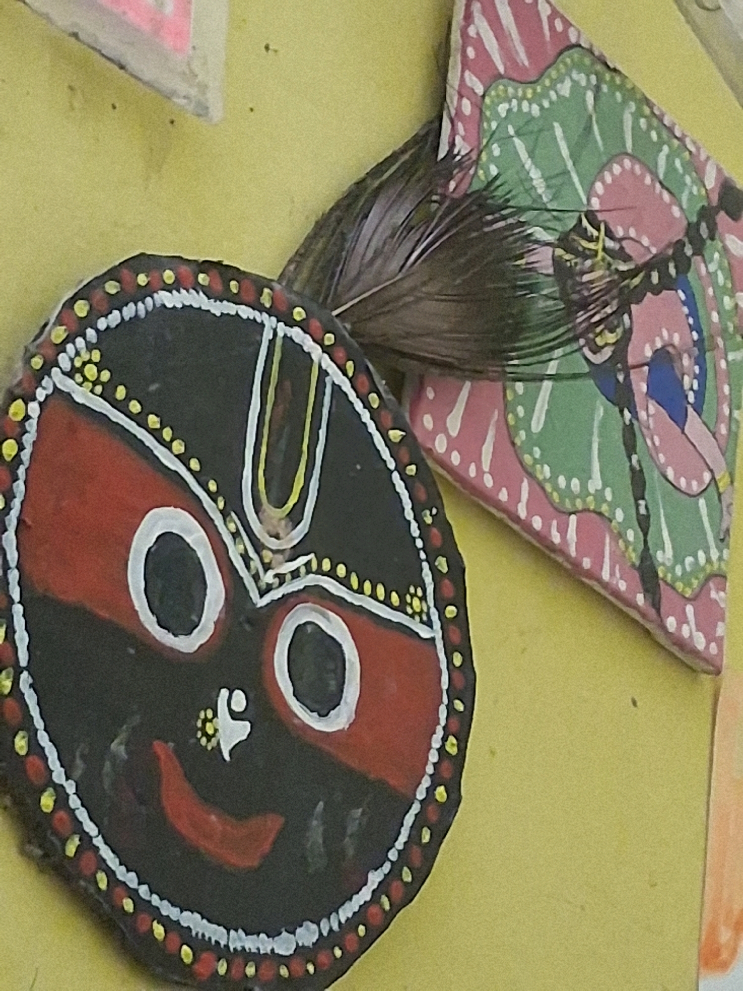 The image displays a close-up view of two decorative items on a pale yellow surface, likely a wall or table. In the foreground and to the left, a circular, hand-painted object depicts a stylized face. This face is primarily black with red markings, two large white-rimmed eyes with black pupils, a small yellow and white depiction of a figure above a red curved mouth. The perimeter of the circle is adorned with a pattern of red and yellow dots separated by white dots. A few dark feathers are positioned on top of this circular object. To the right and slightly behind the circular object, a triangular, hand-painted item features a design with pink and green shapes and white dots, possibly representing a stylized butterfly or floral pattern. The items are displayed in an indoor setting, with the background appearing to be a textured, light yellow surface. The image is captured in Bidhannagar, India.