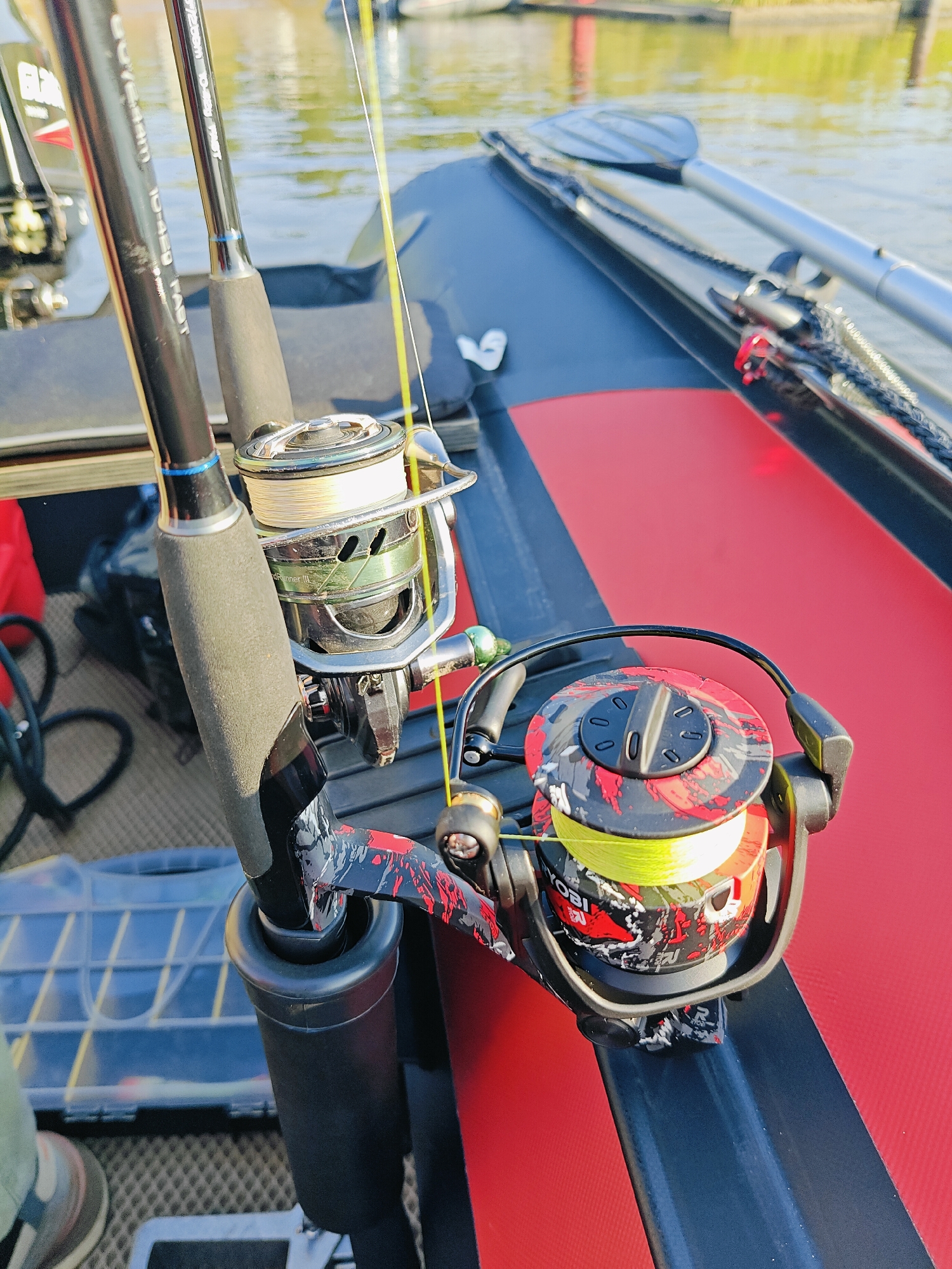 Two fishing rods with reels are mounted on a boat. The boat is dark gray and red, with oars visible in the background. The water appears calm and reflects the sunlight. The fishing rods have black handles and silver/chrome reels. One reel is loaded with light-colored fishing line, while the other has bright yellow line. The scene suggests a fishing trip on a body of water, likely in an urban or suburban setting given the reference to Chaikovsky, Russia, although no specific landmarks are identifiable.