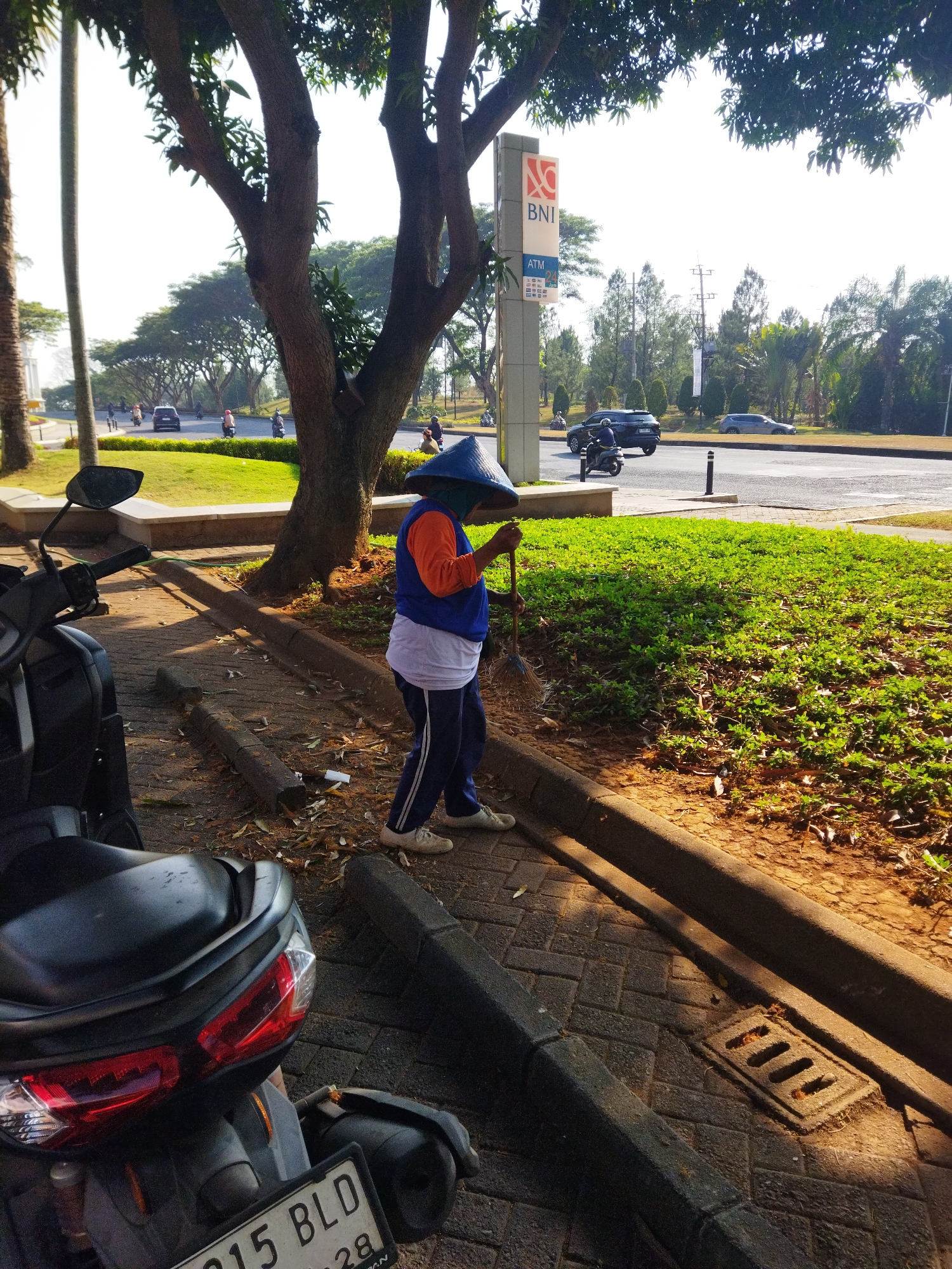 A groundskeeper wearing a conical hat, a blue vest over an orange long-sleeved shirt, and navy blue track pants is sweeping a paved area with a broom. The groundskeeper is positioned to the right of a large tree with a dark trunk. In the foreground, the back of a black motorcycle with a license plate reading "215 BLD" is visible. Behind the groundskeeper, a road with several vehicles and lush green grass can be seen. A BNI bank sign with "ATM 24" displayed on it stands to the left of the tree. The scene appears to be in an urban area, likely on a sunny day, given the shadows. The location is identified as Semarang, Indonesia, implying a tropical or subtropical environment with abundant vegetation.