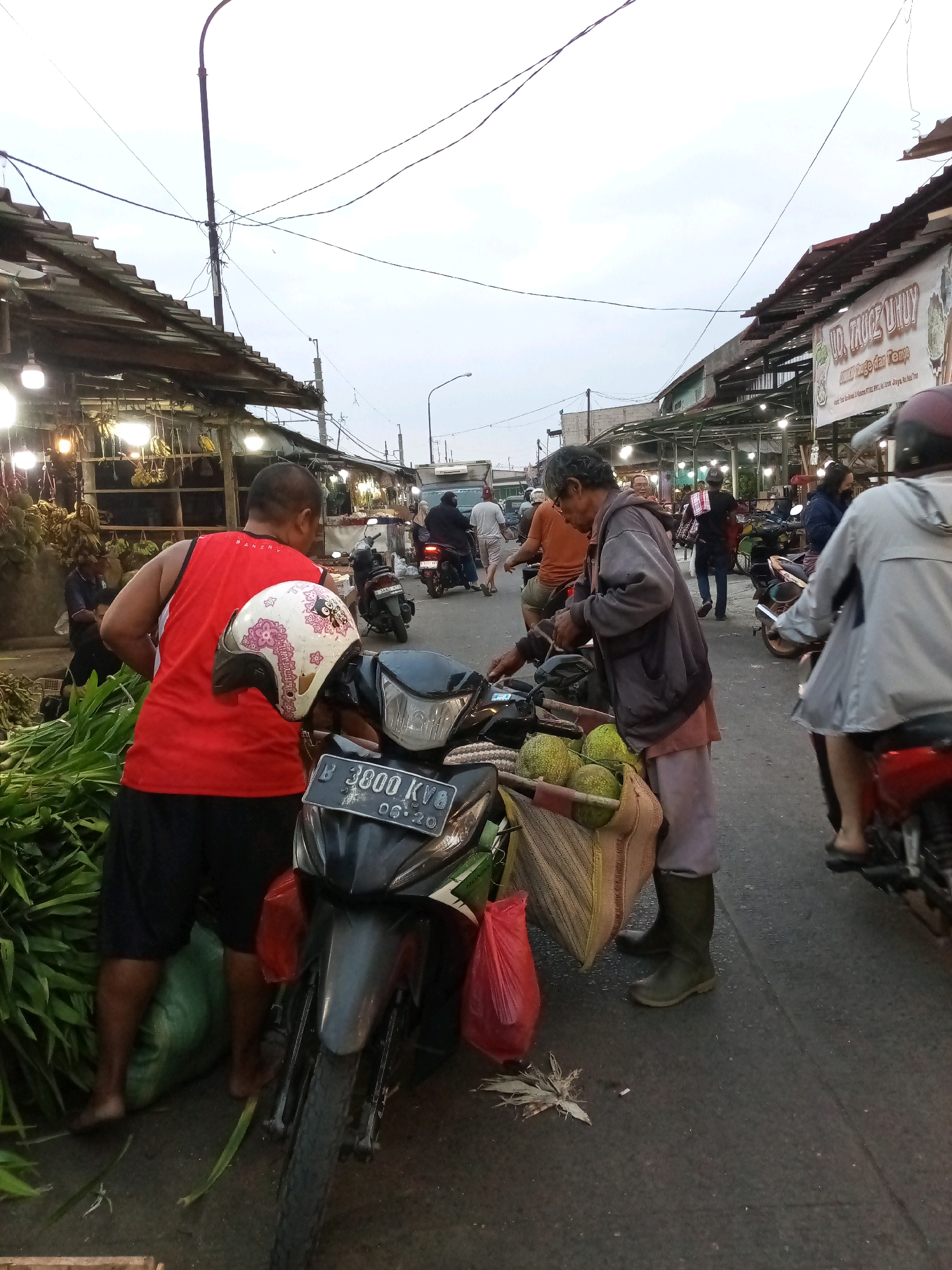 A street market scene is captured in Bekasi, Indonesia, likely in an outdoor marketplace. The image depicts a busy street with several individuals interacting amidst stalls and parked motorcycles. The sky is overcast, suggesting it might be early morning or late afternoon.
In the foreground, a black motorcycle with the license plate "B 3800 KV8" is prominently displayed, with a white helmet adorned with pink floral patterns placed on its handlebars. To the left, a man wearing a red sleeveless shirt and black shorts stands near a large pile of green leafy vegetables. He appears to be interacting with the vegetables, possibly arranging them or preparing them for sale.
To the right of the motorcycle, an older man in a grey jacket and light-colored pants, wearing dark green boots, is leaning over and appears to be examining or handling some produce. He has a basket attached to his motorcycle, filled with round, green fruits, possibly durians. Next to him, a red plastic bag is visible on the ground.
Further into the background, other people are seen walking, standing, and interacting. Several motorcycles are parked along the street. The market stalls on either side of the street have tiled roofs and are lit by overhead lamps. A banner with Indonesian text, including "UD. FROZE UHUK" and "Minuman Rame dan Tempe," is visible on the right side, indicating food stalls. The overall atmosphere suggests a typical Indonesian street market environment, characterized by vibrant activity and commerce.