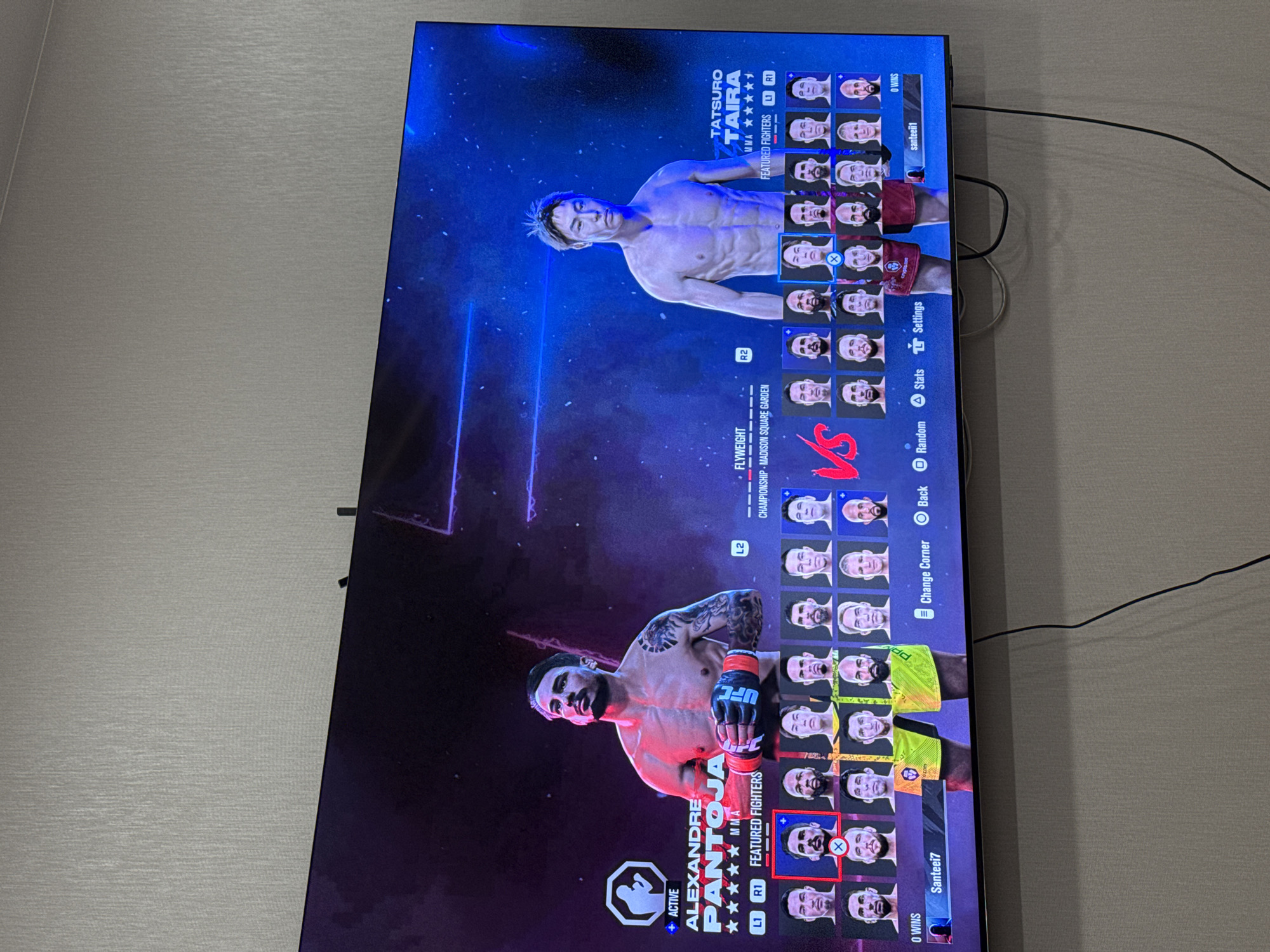 The image shows a television screen displaying a video game featuring mixed martial arts (MMA) fighters. The screen is divided into two sections, each displaying a different fighter: Tatsuro Taira on the top and Alexandre Pantoja on the bottom. Both fighters are shirtless and wearing gloves.
The video game interface shows fighter statistics, player names, and menu options such as "Change Corner," "Back," "Random," "Stats," and "Settings." The game is set in the Flyweight division and appears to be a championship match held at Madison Square Garden.
The television is mounted on a light-colored wall. A black cable is visible near the upper right corner of the screen.