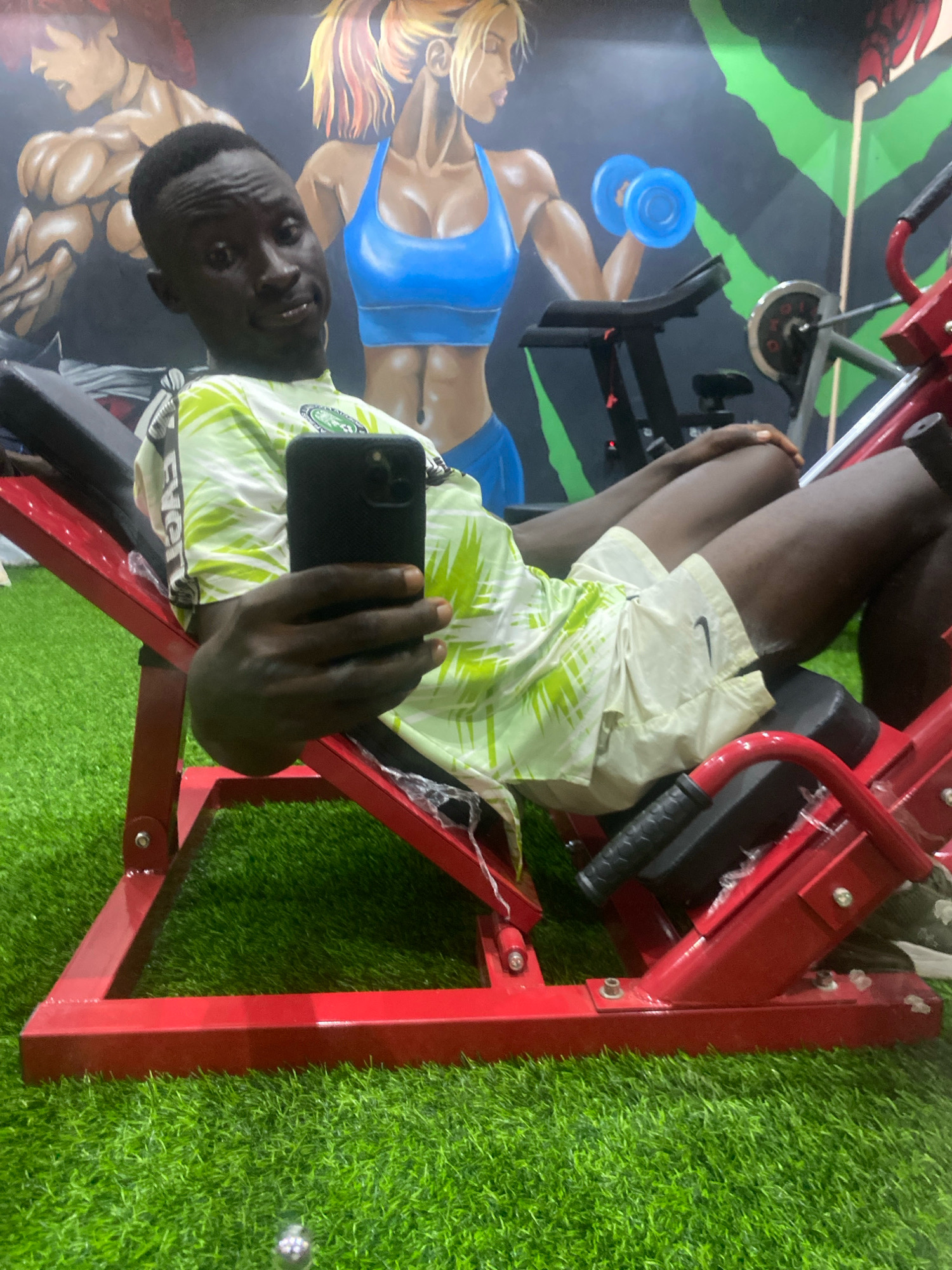 A young man is seated on a red leg press machine in a gym. He is wearing a green and white patterned t-shirt and light-colored shorts. He is holding a black smartphone in his right hand and appears to be taking a selfie. The gym has a vibrant mural on the wall featuring cartoonish depictions of muscular men and women, some holding dumbbells. The floor is covered with artificial green grass. The scene takes place in a gym in Barden Yana, Nigeria.