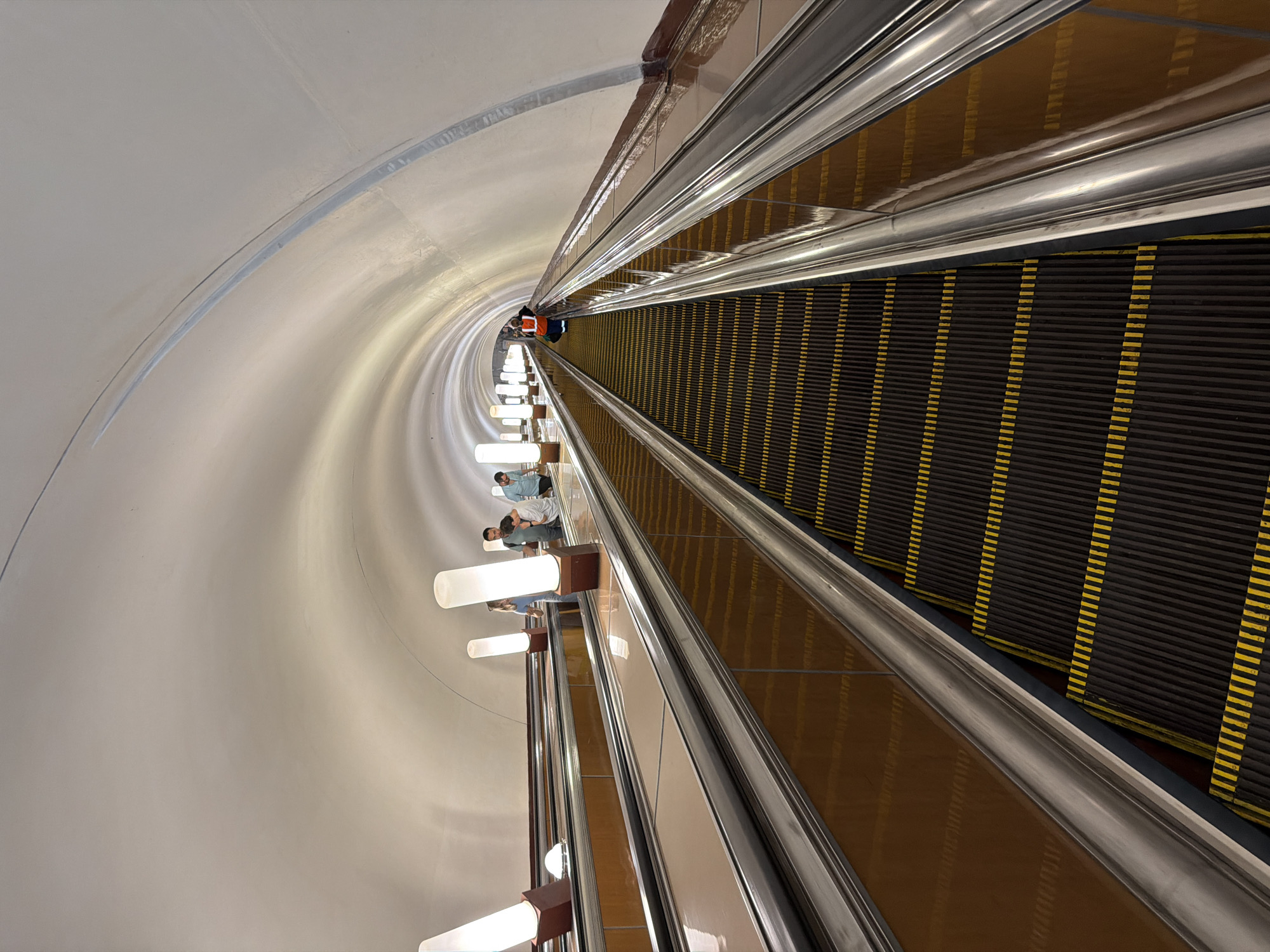 The image shows a view of a Moscow Metro station escalator. The escalator is wide and long, with a curved, white ceiling that extends into the distance. Rows of cylindrical, illuminated lamps are mounted along the sides of the escalator's path, casting a bright, diffused light. The escalator treads are black with yellow safety stripes. The side panels of the escalator are a polished, reflective brown material.
Several people are visible on the escalator. On the left-hand escalator, a woman with blonde hair in a light blue shirt is standing, and to her right, a man and woman in casual clothing are talking. Further up this escalator, another person in a light blue shirt is visible. On the right-hand escalator, a person wearing an orange vest and dark clothing is standing, facing away from the camera. Several other individuals can be seen further down this escalator, appearing to be moving upwards. The overall impression is of a busy public transport hub, with the distinctive architecture of the Moscow Metro.