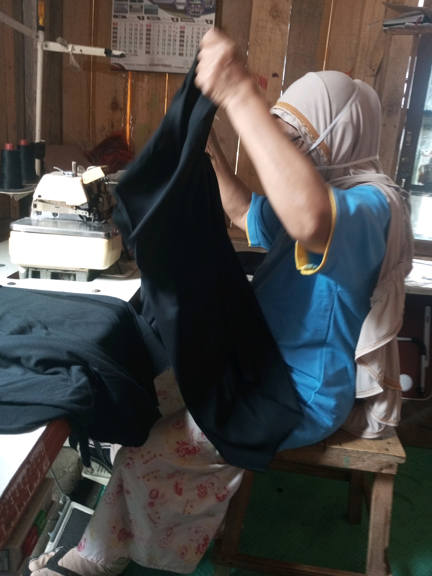 In Kalibeber, Indonesia, a person is seated on a wooden stool in a room with wooden walls. The individual is wearing a light-colored hijab and a blue shirt with yellow trim. They are holding a large piece of dark fabric, possibly for sewing, and a sewing machine is visible in the background. The environment appears to be a workspace, likely a tailor's shop or home studio.