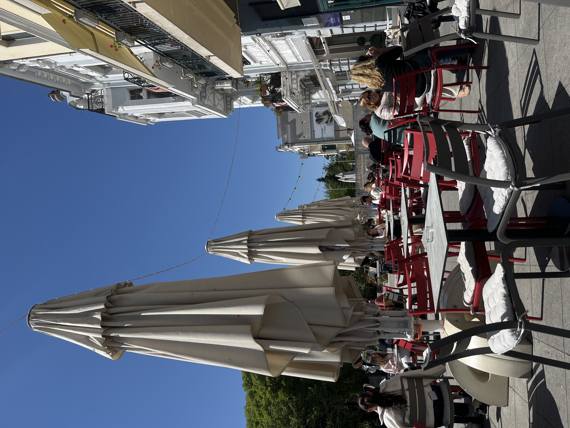 The image shows an outdoor cafe scene in Plovdiv, Bulgaria, under a clear blue sky. Rows of closed, light-colored umbrellas extend along the street, providing shade over the tables and chairs. People are seated at the tables, some appearing to be conversing and others engaged in individual activities. The chairs are primarily red, with a few grey ones present. The buildings lining the street feature ornate architectural details and are generally light in color, with awnings and balconies visible. String lights are strung above the cafe area, adding to the ambiance. The ground is paved.