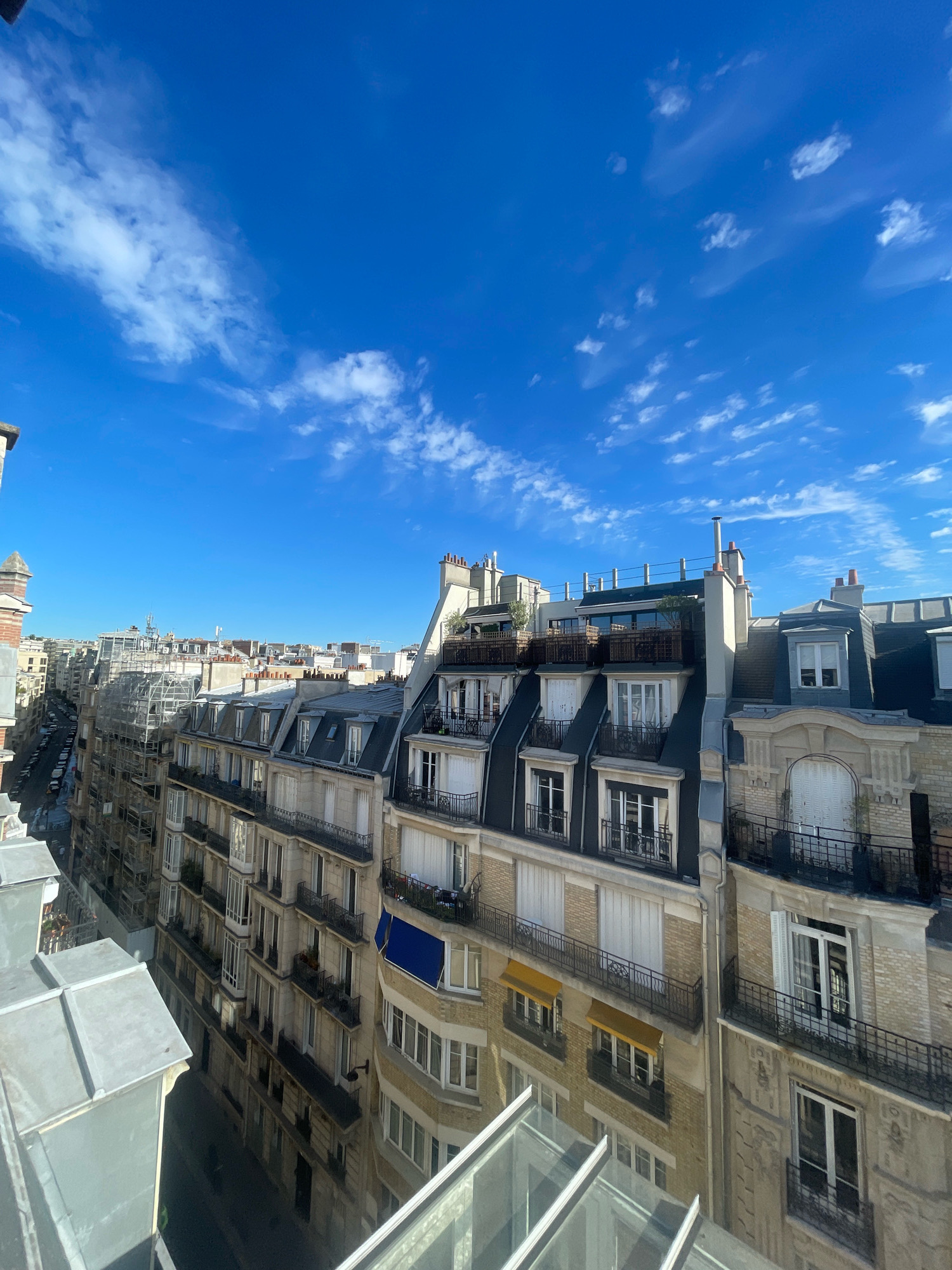 The image depicts a view of Parisian rooftops under a bright blue sky with scattered white clouds. The architecture is characteristic of Paris, with mansard roofs, dormer windows, and stone facades. Several apartment buildings are visible, some with balconies and awnings. In the foreground, a portion of a glass-paneled structure, likely a balcony or solarium, is present. The street below is lined with parked cars, and scaffolding is visible on one of the buildings, suggesting ongoing construction or renovation. The scene captures a typical urban landscape in Paris.