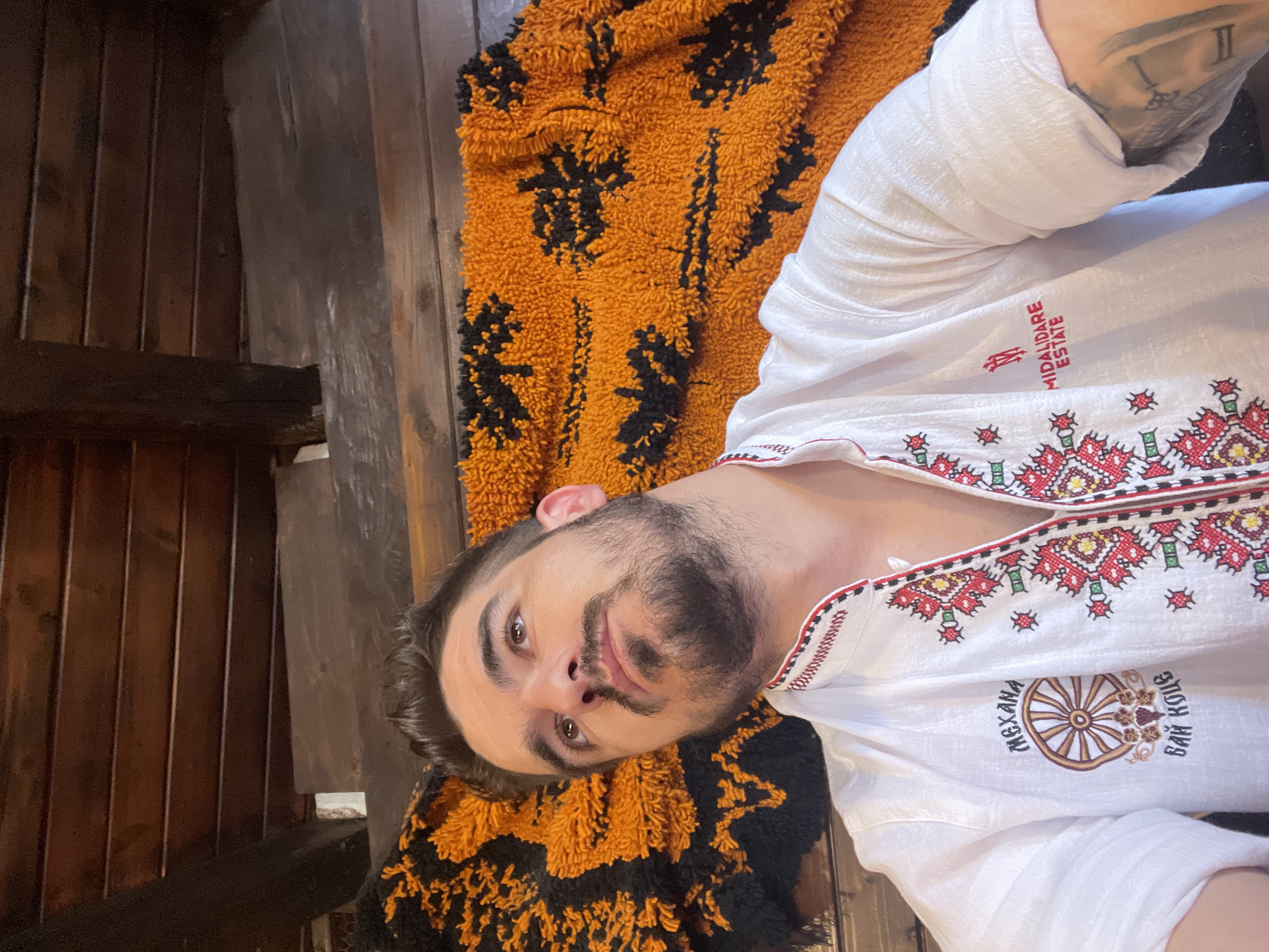 A man with dark hair and a beard is pictured from the chest up, lying down and looking upwards. He is wearing a white shirt with colorful embroidery around the collar and down the front. The shirt has two patches on the left side of the chest, one with a wheel and text, and another with a logo and text. Behind him is an orange and black patterned blanket. The background consists of dark wooden planks. The scene appears to be indoors, possibly in a rustic setting.