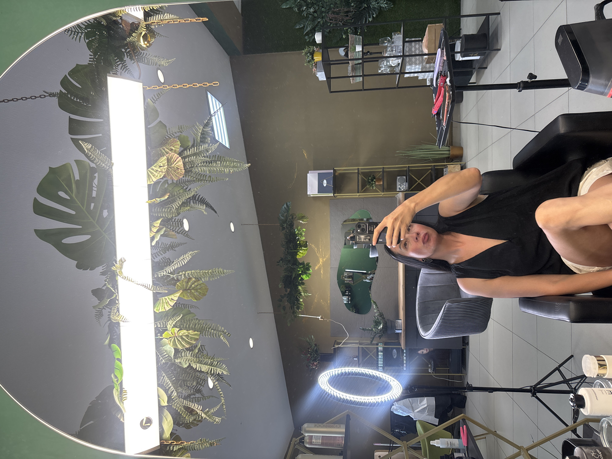 The image captures a woman taking a selfie in a salon. She is seated in a black salon chair, wearing a black sleeveless V-neck top and white shorts. The woman is holding a smartphone in her right hand, angled to take the picture. A ring light is positioned to her left, illuminating the scene. In the reflection of the large mirror behind her, the salon's interior is visible. It appears to be a modern salon with green accents, shelves filled with products, and decorative plants. The overall impression is that of a well-lit and stylish beauty salon. The image is not located in Chaikovsky, Russia.