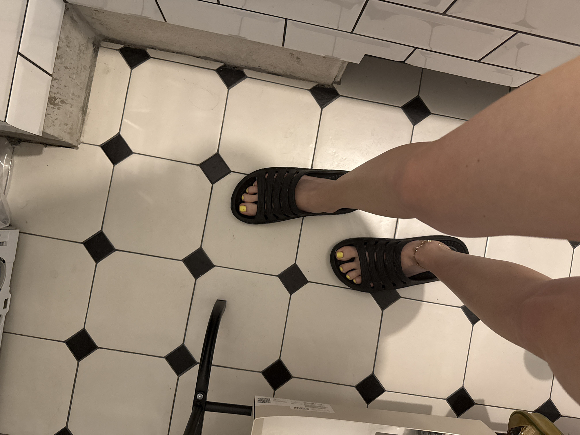 The image shows a person standing on a tiled floor, wearing black sandals with yellow toenail polish. The floor is tiled in a white and black pattern, with a possible step or ledge visible along one edge. Part of what appears to be a chair and an appliance or box are also visible at the bottom of the frame. The location is not discernible from the image. The image does not provide sufficient information to suggest the location is Moscow, Russia.