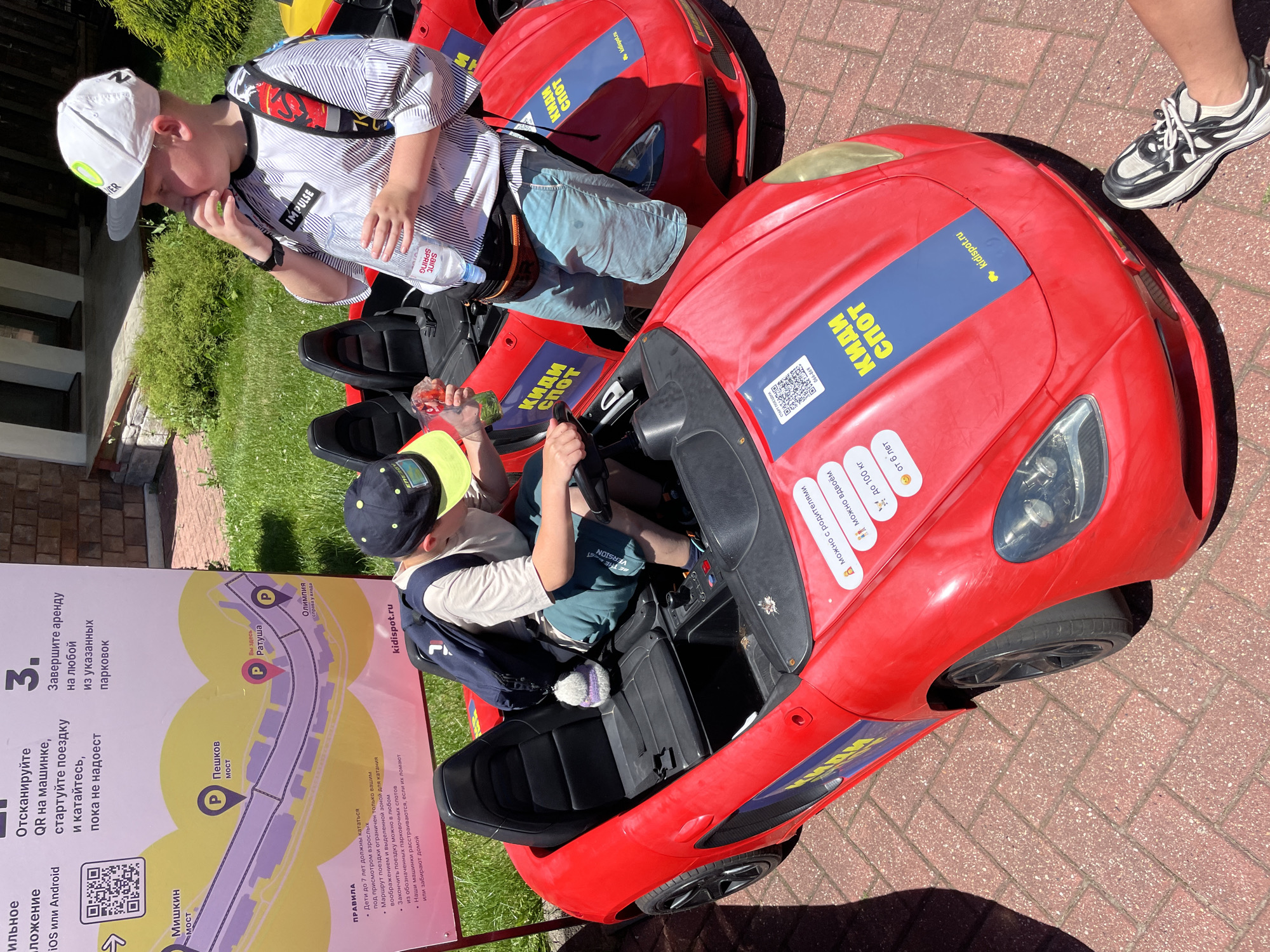 This media file captures a sunny daytime scene in Estosadok, Russia, featuring two young boys in bright red toy cars. The setting appears to be an outdoor recreational area, with green grass and red brick paving stones visible.
The foremost subject is a boy in the driver's seat of a red toy car, wearing a dark baseball cap and a light-colored shirt. He is holding onto the steering wheel, suggesting he is actively engaged in playing with the car. Behind him, another boy is visible, also in a toy car, wearing a white cap and a striped shirt. He is holding a bottle of water and appears to be taking a drink. Both boys are dressed in casual summer clothing, with shorts being visible on the boy in the front car.
The toy cars are bright red and have stickers with blue accents. One sticker on the car in the foreground displays the text "КИДИ СПОТ" and a website address "kidispot.ru" with a QR code. Another sticker reads "можно с родителями" (possible with parents).
To the left of the scene, a large vertical sign with a map and text in Russian provides context. The map shows different locations within a park or recreational area, with icons indicating parking spots and names like "Пешков Мост" (Peshkov Bridge) and "Олимпиада" (Olympia). The text on the sign explains rules and instructions for using the toy cars, including scanning a QR code to start the rental, information about the age and weight limits for children, and rules about driving and returning the cars.
The overall impression is one of a fun, family-friendly outdoor activity on a warm day. The sunlight creates shadows, indicating it's likely midday.