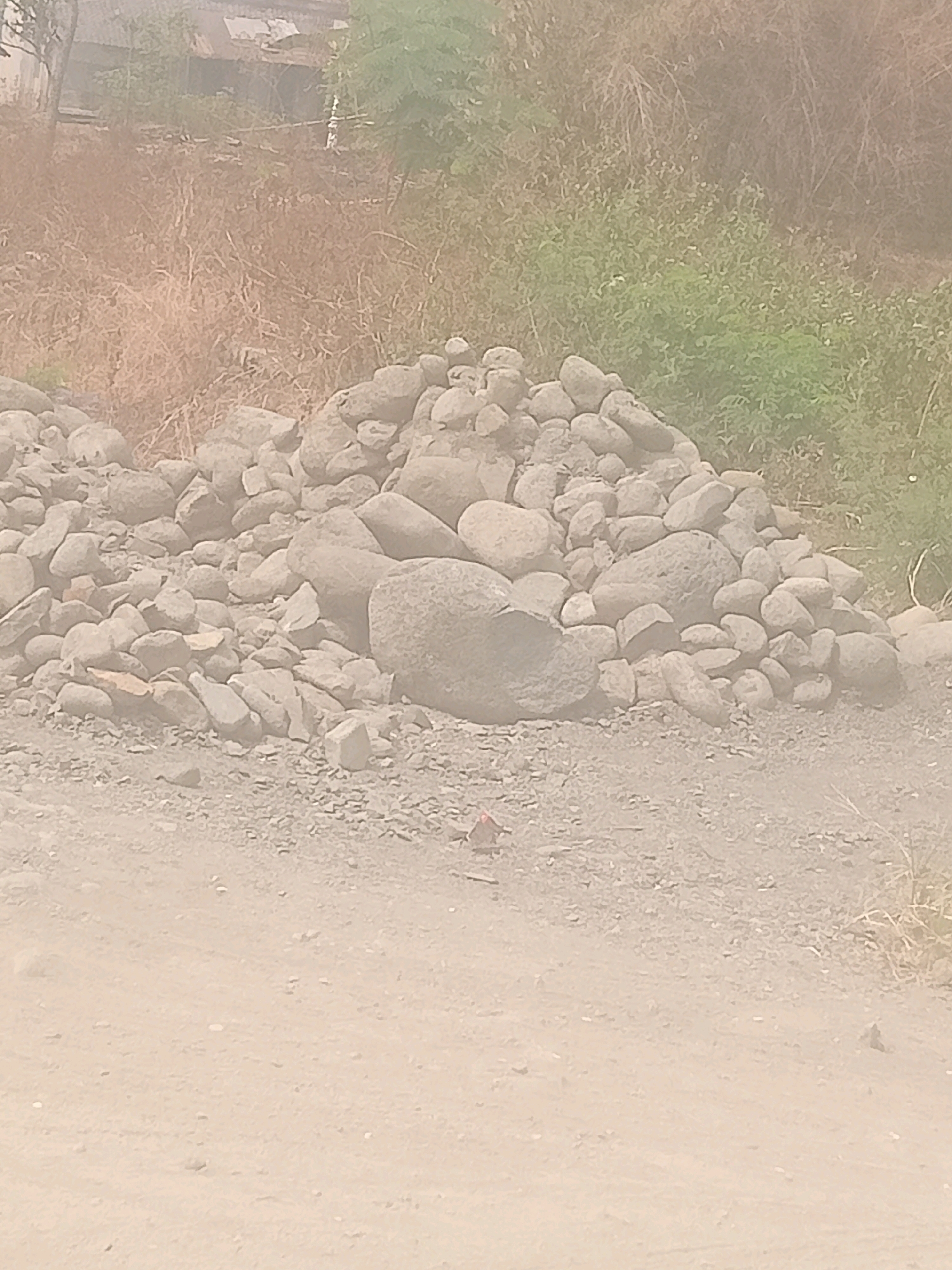 In Pendawa, Indonesia, a pile of large, rounded river stones is situated on a dusty, gravelly embankment. Behind the stones, dry, brown vegetation and a cluster of green bushes are visible, suggesting a semi-arid environment. The background shows what appears to be a simple structure, possibly a dwelling, with a corrugated roof, partially obscured by trees. A small, red object, potentially a piece of litter or debris, is located on the ground in front of the stone pile. The lighting is bright, indicating daytime, and the overall scene is one of a rural or undeveloped area.