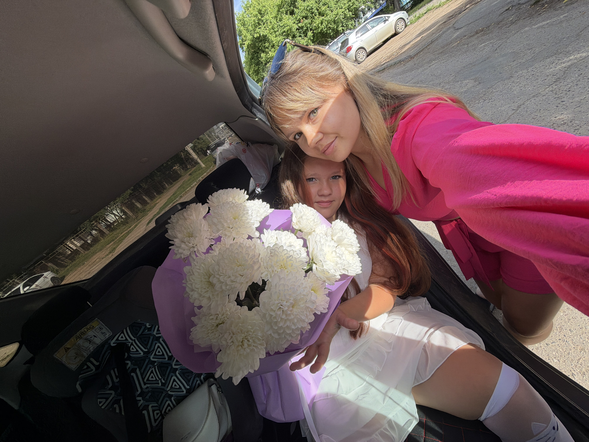 In Chaikovsky, Russia, a young woman with long blonde hair, wearing a bright pink dress, is taking a selfie with a young girl. The girl is seated in the trunk of a car, wearing a white dress and white stockings adorned with cat faces. She is holding a large bouquet of white chrysanthemums wrapped in light purple paper. The car appears to be parked on a paved surface, possibly a street or parking lot, with trees and other vehicles visible in the background. The lighting suggests it is daytime, with sunlight casting shadows. The trunk of the car is open, revealing the interior and a car seat on the left side. The overall scene suggests a celebratory or gifting occasion.