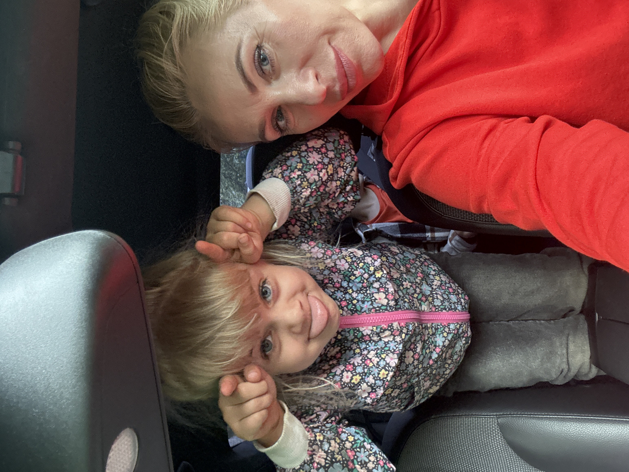 A selfie is taken inside a car, likely in Saint Petersburg, Russia, based on the inferred location. In the foreground, a woman with blonde hair pulled back smiles at the camera. She is wearing a bright red hooded sweatshirt. To her left, a young child with blonde hair is making a funny face with their tongue sticking out and fingers pointed towards the camera. The child is wearing a colorful floral jacket with a pink zipper over grey pants. The background shows the interior of the car, including car seats and the ceiling console.