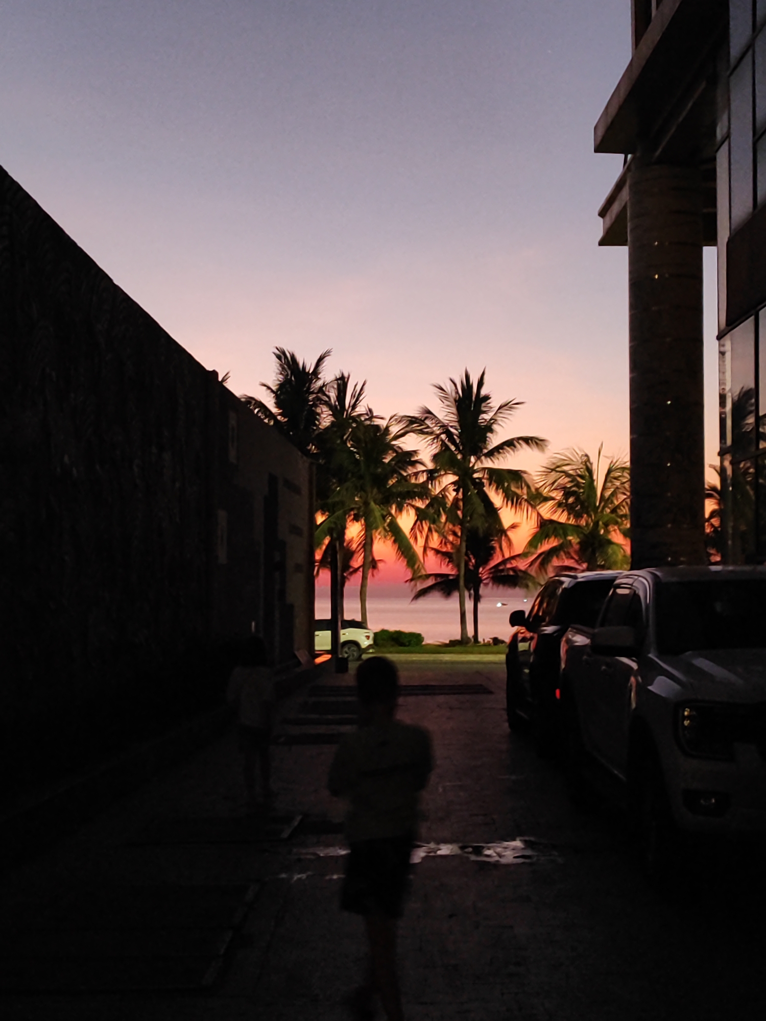 The image shows a street scene in Đà Nẵng, Vietnam, at sunset. The sky is a gradient of purple, pink, and orange. Palm trees are silhouetted against the bright sky in the distance. Two children, partially obscured by darkness, are walking on the street. Two pickup trucks are parked along the right side of the street, and a white car is visible in the distance. Buildings frame both sides of the street. The scene is dimly lit due to the time of day and the shade cast by the buildings. The street surface appears wet.