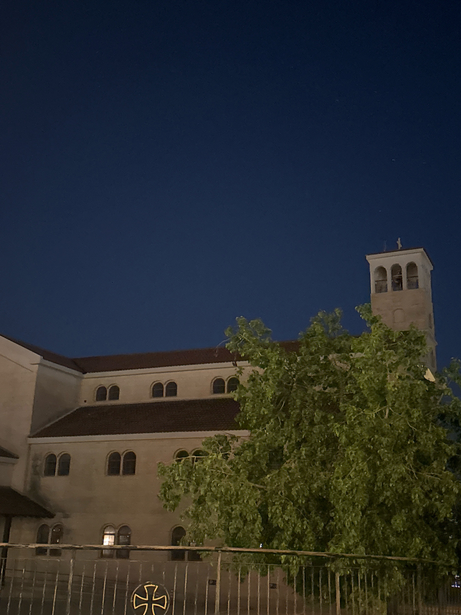 An exterior nighttime photograph showcases a large building with a bell tower against a dark blue sky. The building appears to be a church or similar religious institution, constructed of light-colored stone with a tiled roof. Multiple arched windows are visible on the facade. A mature tree with dense green foliage partially obscures the building and the tower. In the foreground, a metal fence with vertical bars runs horizontally across the bottom of the frame. A decorative cross emblem is visible on the fence. The scene is set in Germasogeia Municipality, Cyprus.