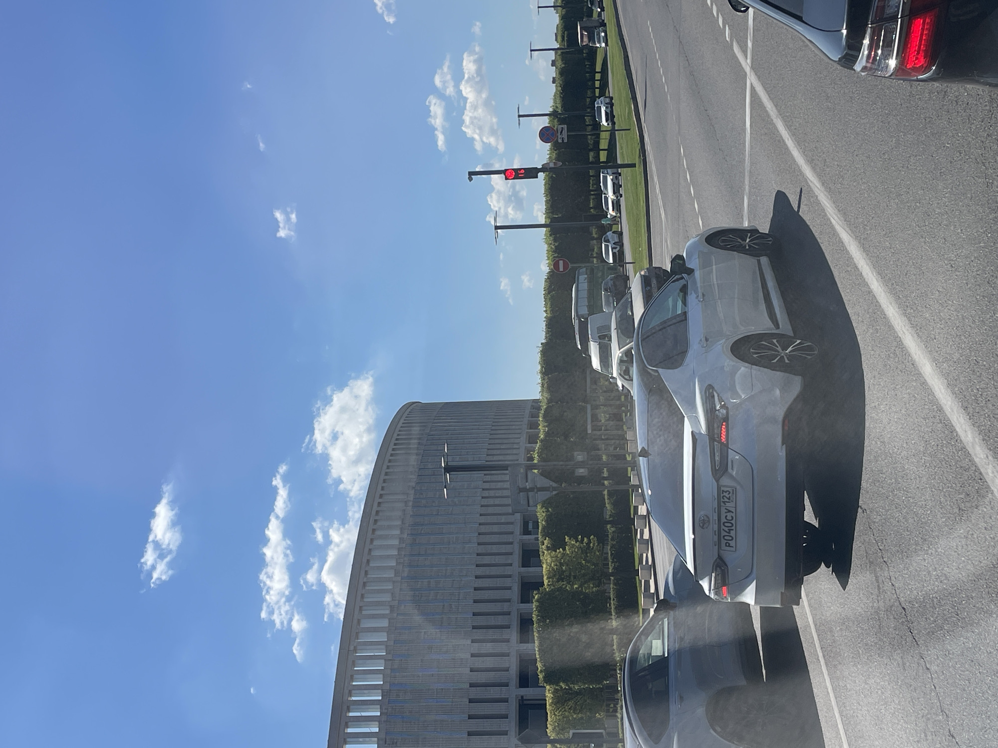 The image depicts a street scene in Krasnodar, Russia, likely in the vicinity of the FC Krasnodar Stadium, given the architectural style of the large, curved building in the background. The day is sunny with scattered white clouds in a bright blue sky. Several cars are visible on the asphalt road, including a white Toyota Camry in the foreground with the license plate "P040CY 123". A dark-colored sedan is partially visible in the lower right corner. Other vehicles, including a white van and a truck, are present further down the road. A traffic light displaying a red signal is visible on the right side of the image. The road is marked with white lines. Lush green trees and manicured hedges line the side of the road, separating it from the large, modern building.