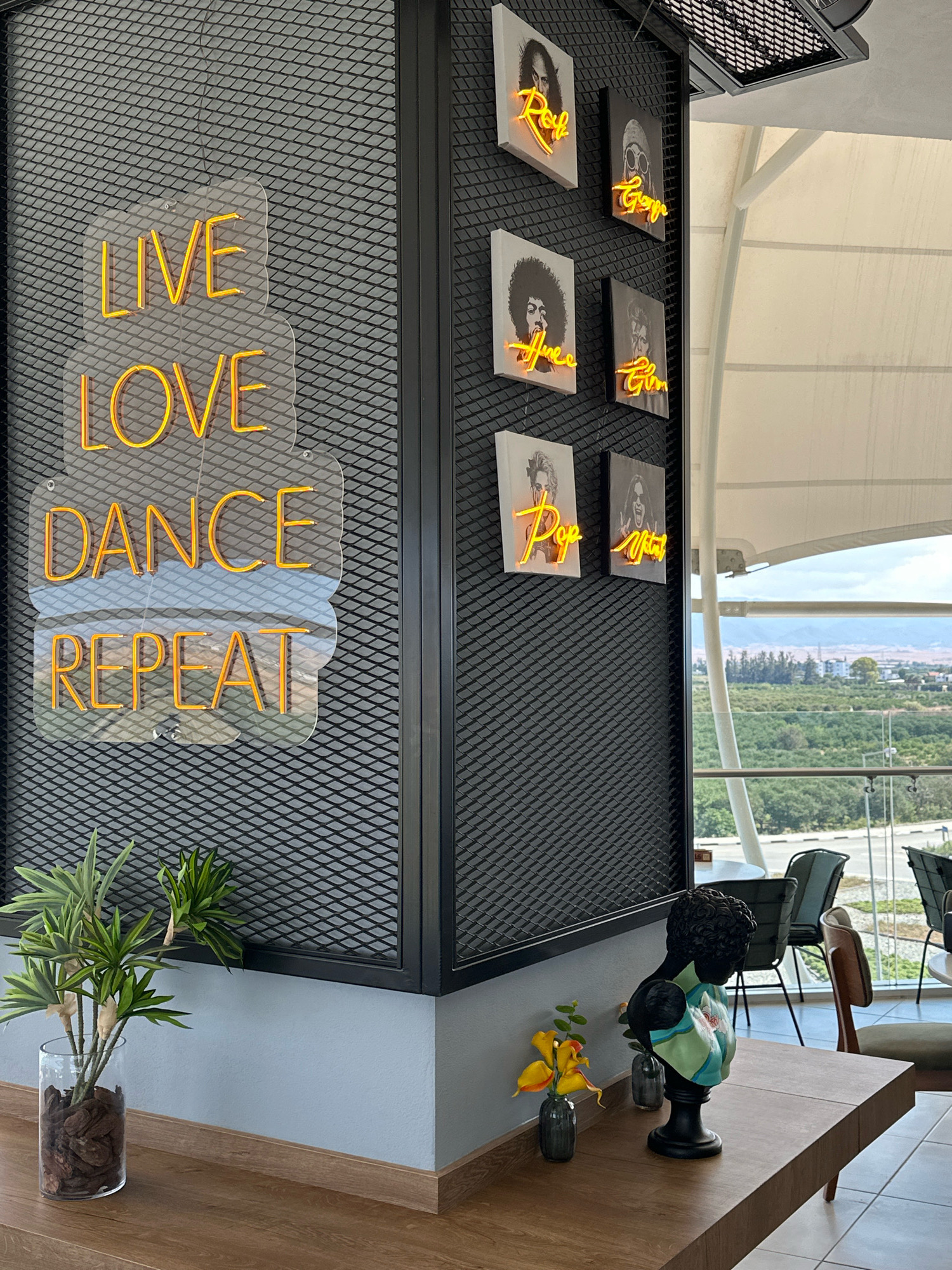In the city of Guzelyurt, Northern Cyprus, an interior scene depicts a wall adorned with yellow neon signs spelling "LIVE," "LOVE," "DANCE," and "REPEAT." These signs are mounted on a clear acrylic backing against a black diamond-patterned metal mesh. To the right of the neon signs, a series of framed pictures featuring portraits with neon script are arranged vertically on the same mesh wall. In the foreground, a wooden surface holds a glass vase with a green plant, and further to the right, a bust with a patterned scarf sits next to small vases and a yellow artificial flower. Beyond the interior, a large window offers a view of a landscape with trees and distant buildings under a cloudy sky, suggesting a balcony or outdoor seating area.