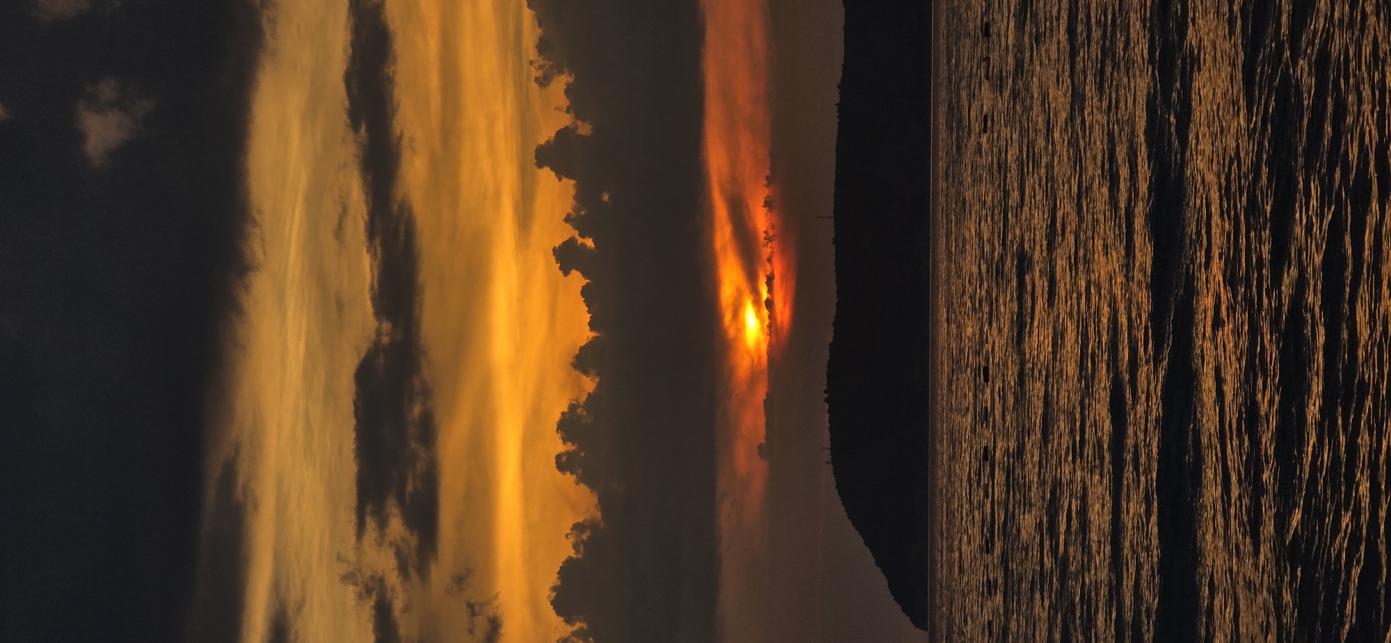 This media file depicts a sunset over a body of water, likely the Adriatic Sea, with the coast of Bar, Montenegro visible in the background. The scene is split vertically, with the left side featuring a dramatic sky and the right side showing the rippling water.
The sky is ablaze with intense oranges and yellows, indicative of a sunset. Streaks of dark clouds are interspersed with the vibrant hues, creating a sense of depth and contrast. The sun itself is partially obscured by clouds, emitting a bright, fiery glow. In the distance, a dark, silhouetted landmass, presumably the coastline, forms the horizon line. A few faint, vertical structures, possibly masts or antennas, can be discerned on the distant land.
The right half of the image is dominated by the texture of the water's surface. The setting sun casts a warm, golden light that reflects off the ripples, creating a pattern of bright lines against a darker background. The water appears to be calm but textured, suggesting a gentle breeze.
The overall mood is one of natural beauty and tranquility, with the vibrant colors of the sunset evoking a sense of awe. The time of day is clearly sunset, and the location cues suggest a coastal environment. There is no visible text in the media file.