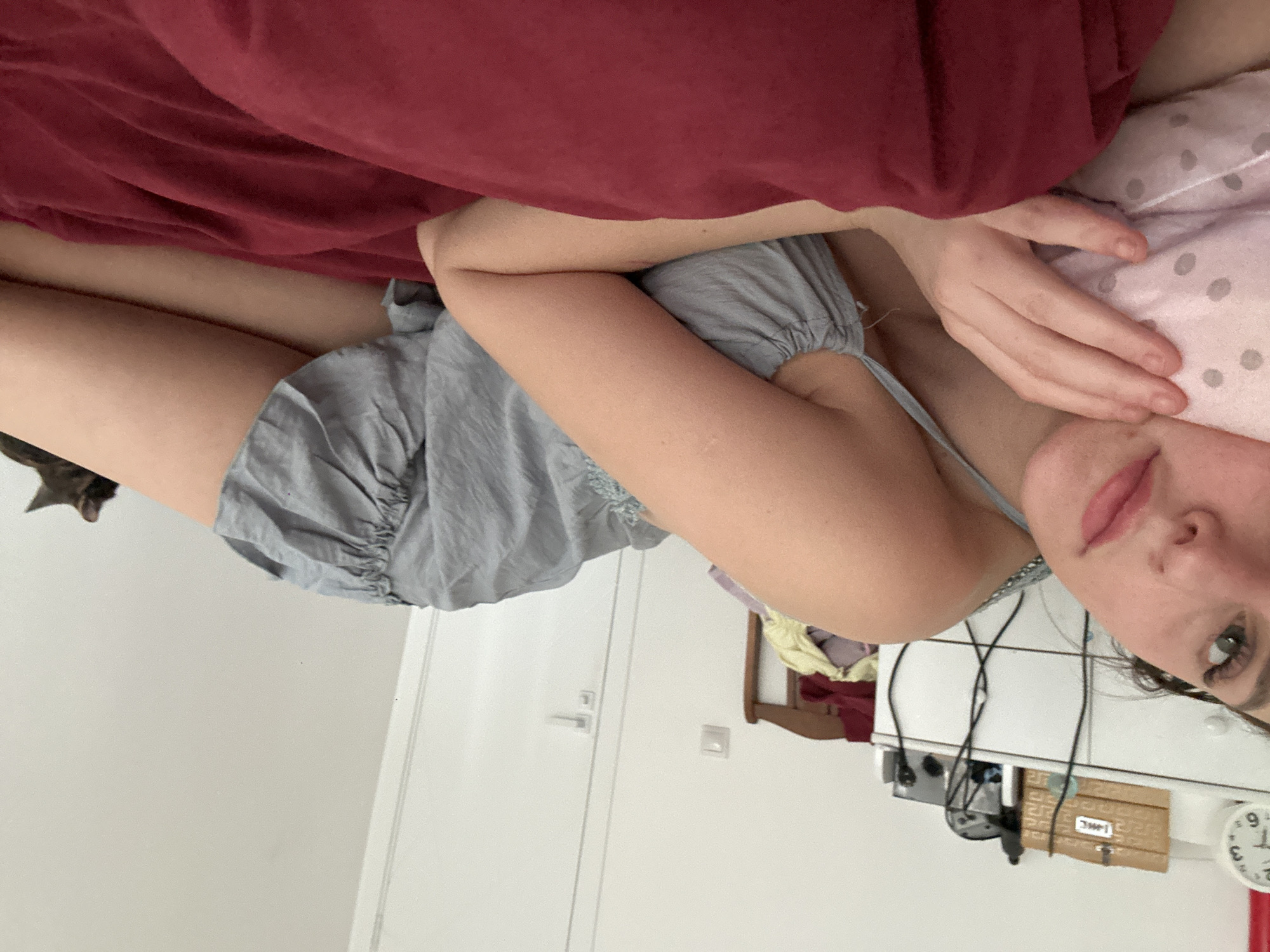 A young woman is lying on a bed, with her head resting on a pink polka-dotted pillow. She is wearing a light blue ruffled dress. Her right arm is bent at the elbow, and her hand is resting on the pillow. Her left arm is out of view. The bed is covered with a dark red comforter. In the background, a white dresser is visible, with various items on top, including a clock and a wooden box. A partially visible cat is lying on the bed in the background to the right. The room appears to be a bedroom in a residential setting.