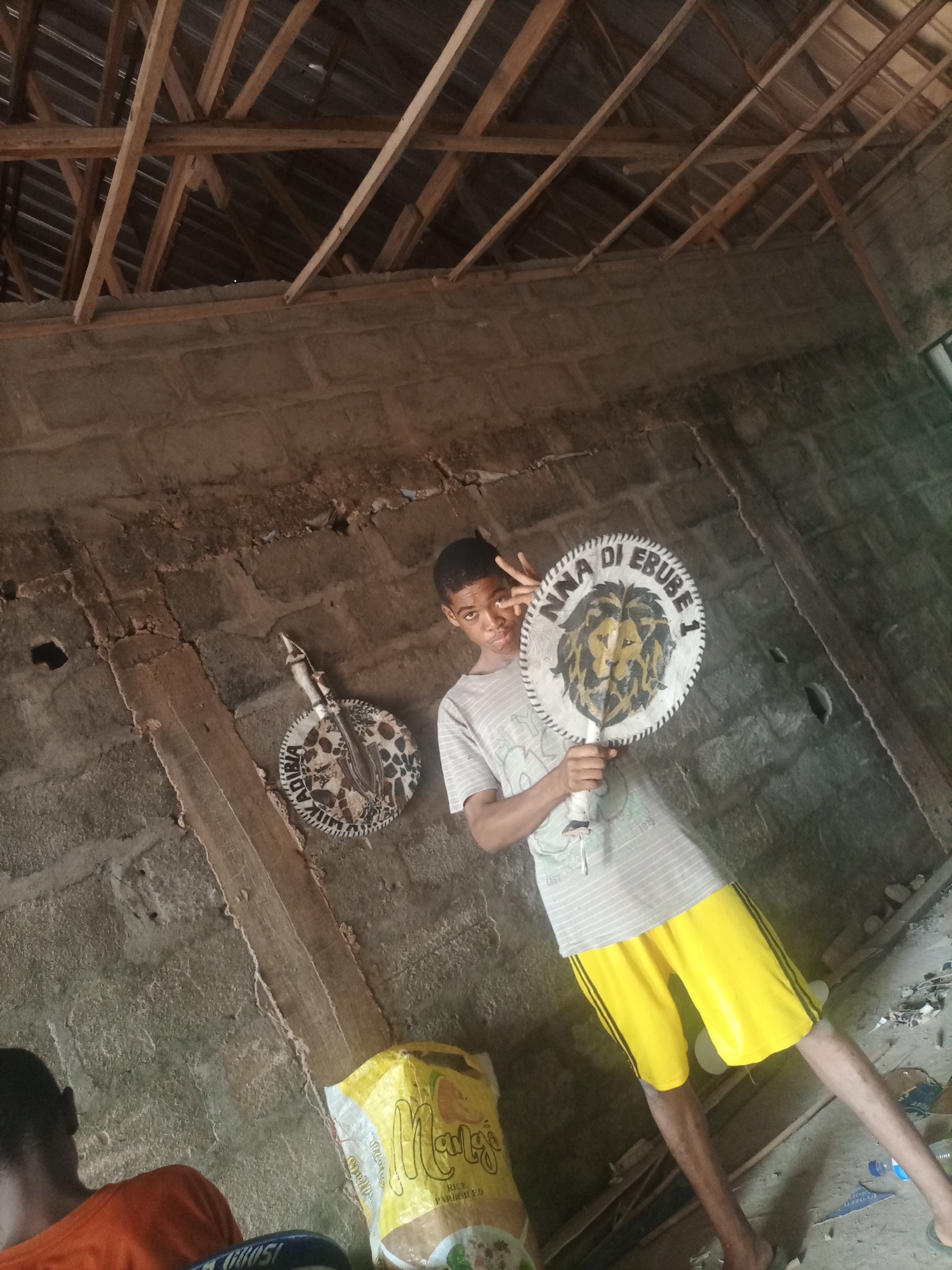 In this photo, a young person is posing in an unfinished room, holding a circular object with a lion design and text that reads "NNA DI EBUBE 1." Another person is visible in the lower left corner, wearing an orange shirt. The room has a concrete block wall and a wooden roof structure that is incomplete. Debris is scattered on the floor, suggesting construction or disuse. A bag with text that appears to be "Maltes" is also visible on the floor. The lighting suggests it might be daytime, but there are no clear indicators of the weather or specific time. The overall atmosphere is casual and perhaps a bit unkempt, with the young person's pose appearing playful.