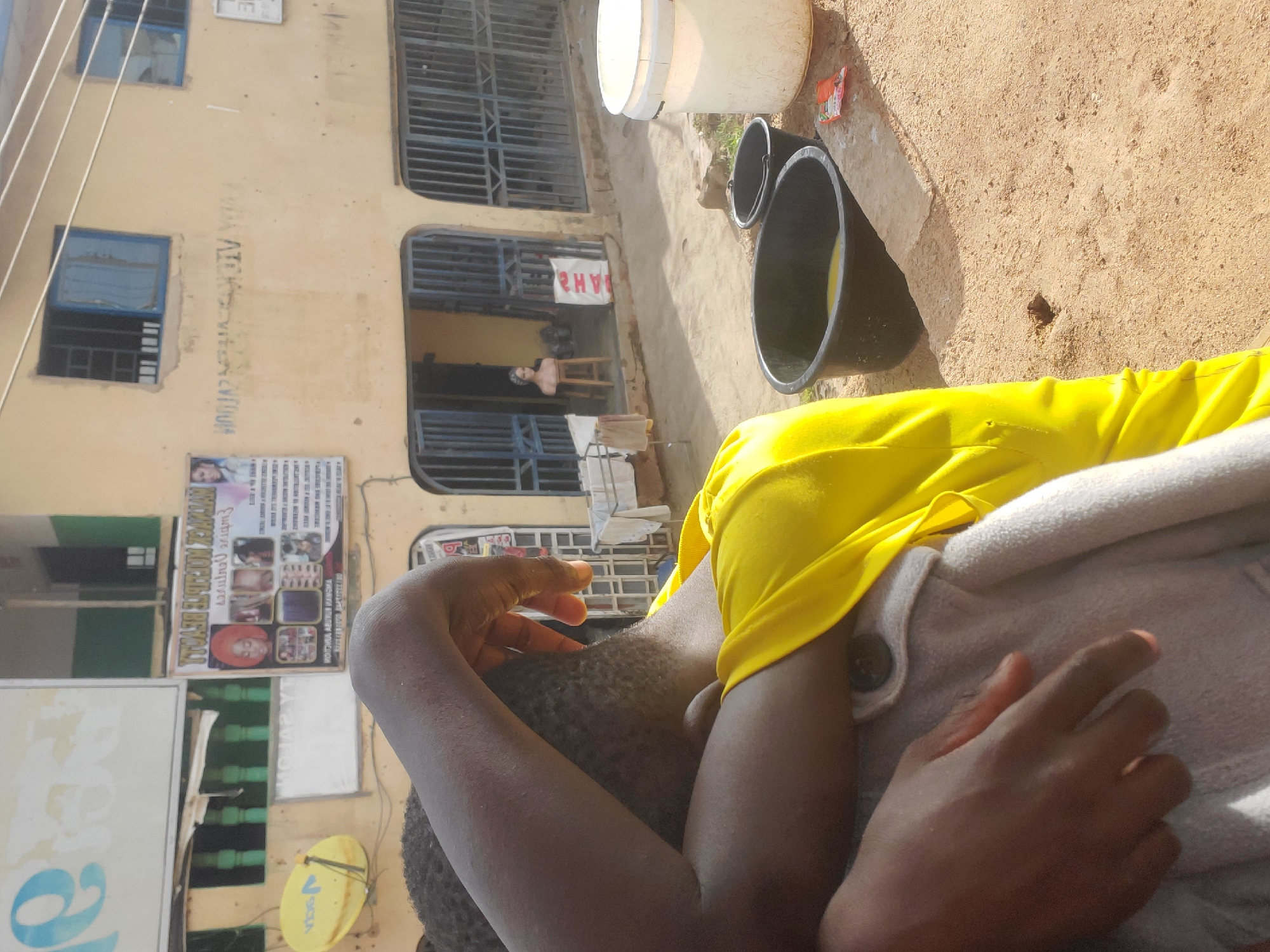 The image depicts an outdoor scene in Gwong, Nigeria. The primary subjects are a person, partially visible and dressed in a yellow shirt and gray outerwear, and several objects. The setting appears to be a dusty, unpaved area next to a weathered building. The building has a light yellow facade and features barred windows and doorways. Some windows have blue panes.
Visible objects include two black buckets and a larger white bucket. A small, colorful packet, possibly food or candy, rests on the ground near the buckets. The person's hands are prominent, with one resting on their head and the other on their lap. Their skin tone is dark, and their hair is short and textured.
The image composition suggests a casual, perhaps mundane moment. The sunlight hitting the ground indicates it is daytime, likely in the morning or afternoon, casting shadows and highlighting the textures of the environment. There is no specific event clearly taking place, but the presence of buckets might suggest preparations for water collection or storage.
A sign is affixed to the building, featuring text and images related to hair styling, with a woman's face prominently displayed. While much of the text on the sign is difficult to read due to resolution and orientation, some words appear to be in English, with "HAIR STYLE" potentially visible. Another sign with large blue lettering is partially visible on the left. A satellite dish is also mounted on the building. The overall impression is one of a typical day in a developing urban or semi-urban area in Nigeria.