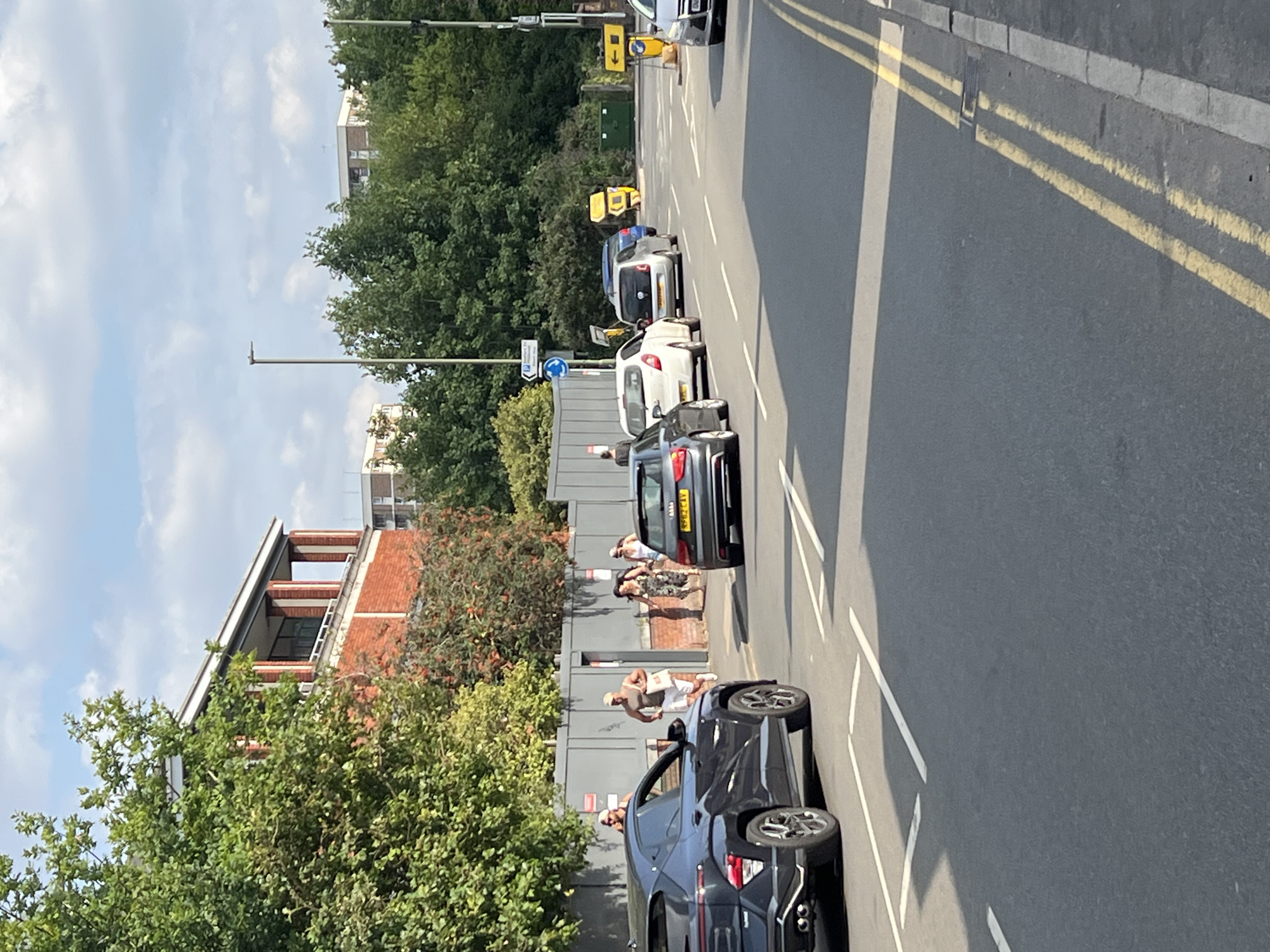The image depicts a street scene in Guildford, United Kingdom, on a sunny day with scattered clouds. In the foreground, a dark SUV is parked on the left side of the road. Several other cars are visible in a queue on the road, including a silver Audi, a white hatchback, and a silver hatchback.
On the right side of the road, a few individuals are walking on a sidewalk. In the background, there is a brick building with a colonnaded upper level, flanked by trees and hedges. Beyond this, taller buildings are visible in the distance. The street is marked with white lines, and double yellow lines run along the curb on the right side. Traffic lights and signage are also present, including a blue circular directional sign. The overall atmosphere is one of urban activity on a bright day.