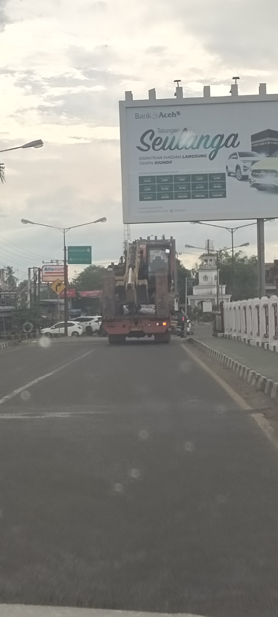 The image captures a road scene from a vehicle, with a large flatbed truck carrying an excavator in the immediate foreground, moving away from the viewer. The road appears damp or reflective, possibly due to rain or light conditions, and there are visible specks on what could be a windshield or camera lens.
Above the road, under an overcast sky, a large billboard prominently displays an advertisement for "Bank Aceh," featuring "Tabungan Seulanga" (Seulanga Savings). The billboard promises "DAPATKAN HADIAH LANGSUNG TANPA DIUNDI!!" (GET DIRECT PRIZES WITHOUT A DRAW!!) and includes a table with various monetary values and images of cars, likely as prizes. The bank's website, "www.bankaceh.co.id," is also visible.
To the left, streetlights illuminate the area, and a green road sign with text such as "WEATANG RAYA" provides directional information. Below it, a yellow road sign indicates a curve. An "Alfamart" convenience store sign can be seen further down the street. Several other vehicles, including white cars, are visible in traffic. On the right side of the road, a white decorative barrier lines the street, and a distinctive white architectural structure resembling a mosque with a clock tower is visible in the distance. The scene suggests a cloudy daytime setting in an urban or semi-urban area, consistent with Dayahlhoksukon Barat, Indonesia.