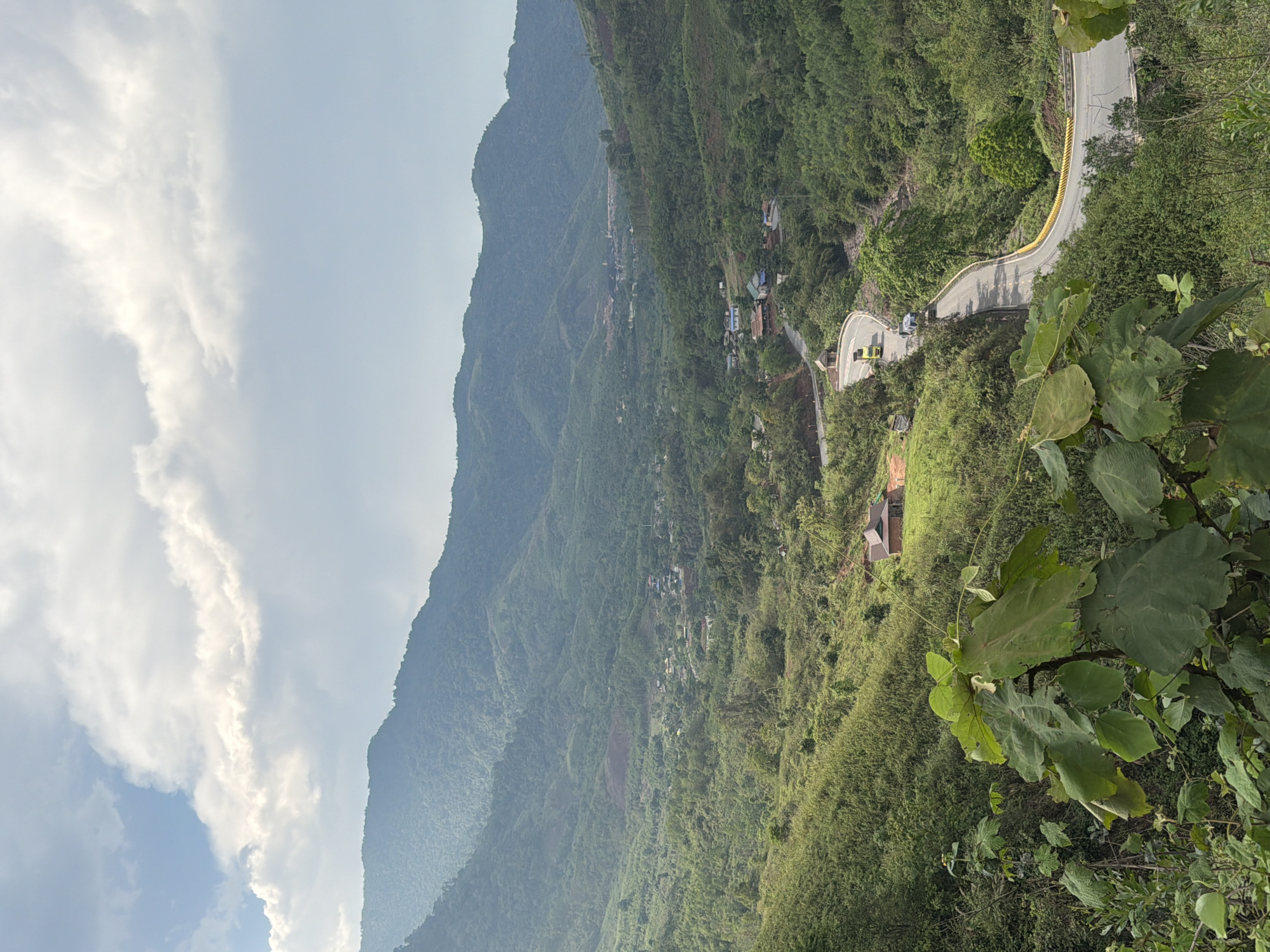 The image depicts a mountainous landscape in La Lay Commune, Vietnam. A winding road cuts through the densely vegetated terrain, with yellow guardrails visible along the edge. A small village is nestled on the side of the mountain, marked by scattered houses. The sky is partly cloudy. In the foreground, large green leaves obscure a portion of the view. Two cars are driving on the road.