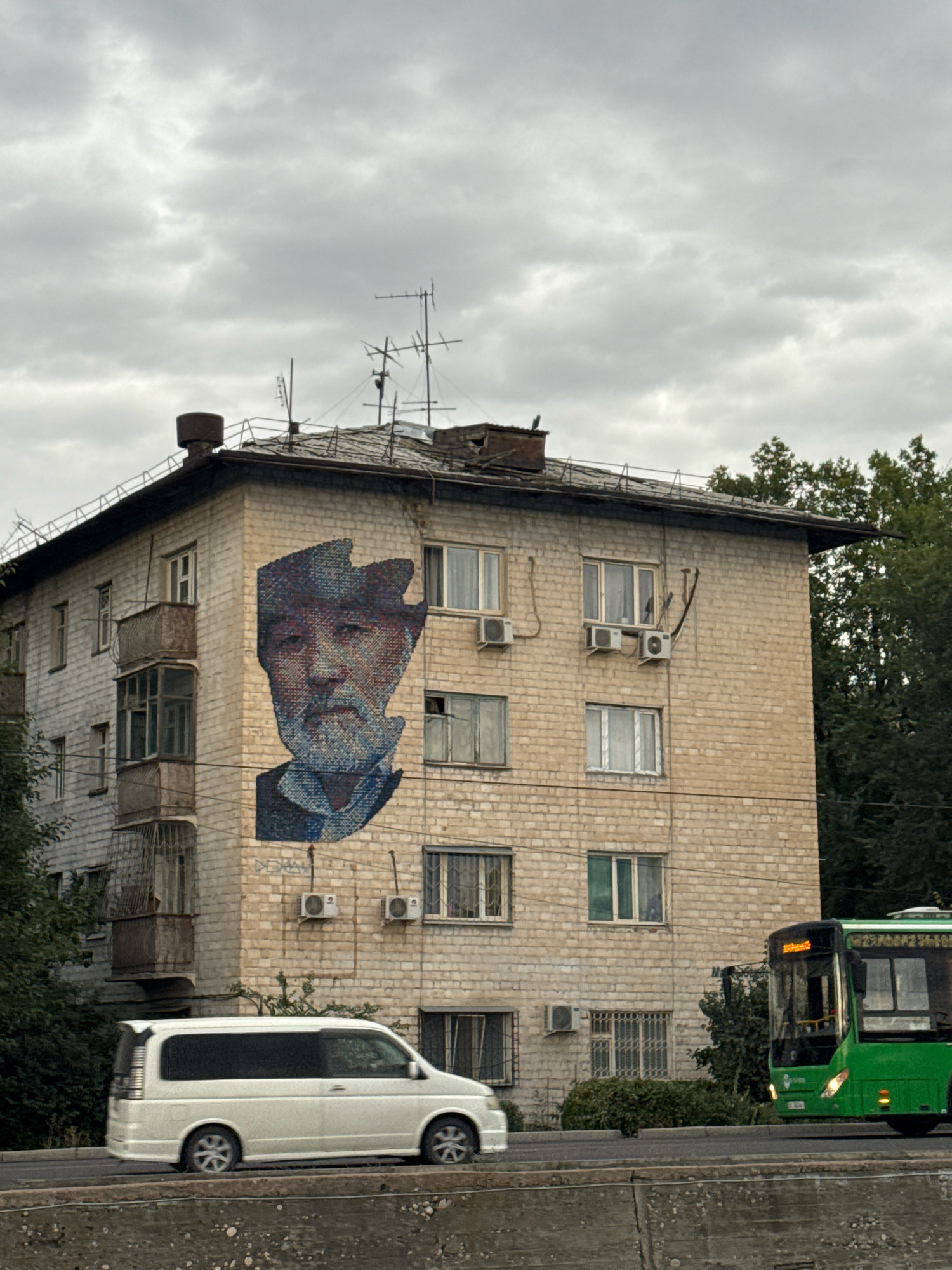 A mural depicting the face of an elderly man with a beard and wearing a hat is painted on the side of a light-colored brick apartment building. The mural is rendered in a mosaic style with small, square tiles in shades of blue, red, and white. The building has multiple windows, some with balconies and some with protective bars. Air conditioning units are visible on the exterior walls. In front of the building, a white minivan is driving on a road. To the right of the building, a green bus is also visible on the road, with its headlights on. Trees and foliage are present in the background. The sky is overcast with gray clouds. The scene appears to be in an urban environment, likely Bishkek City, Kyrgyzstan, based on the architectural style and the presence of public transportation.