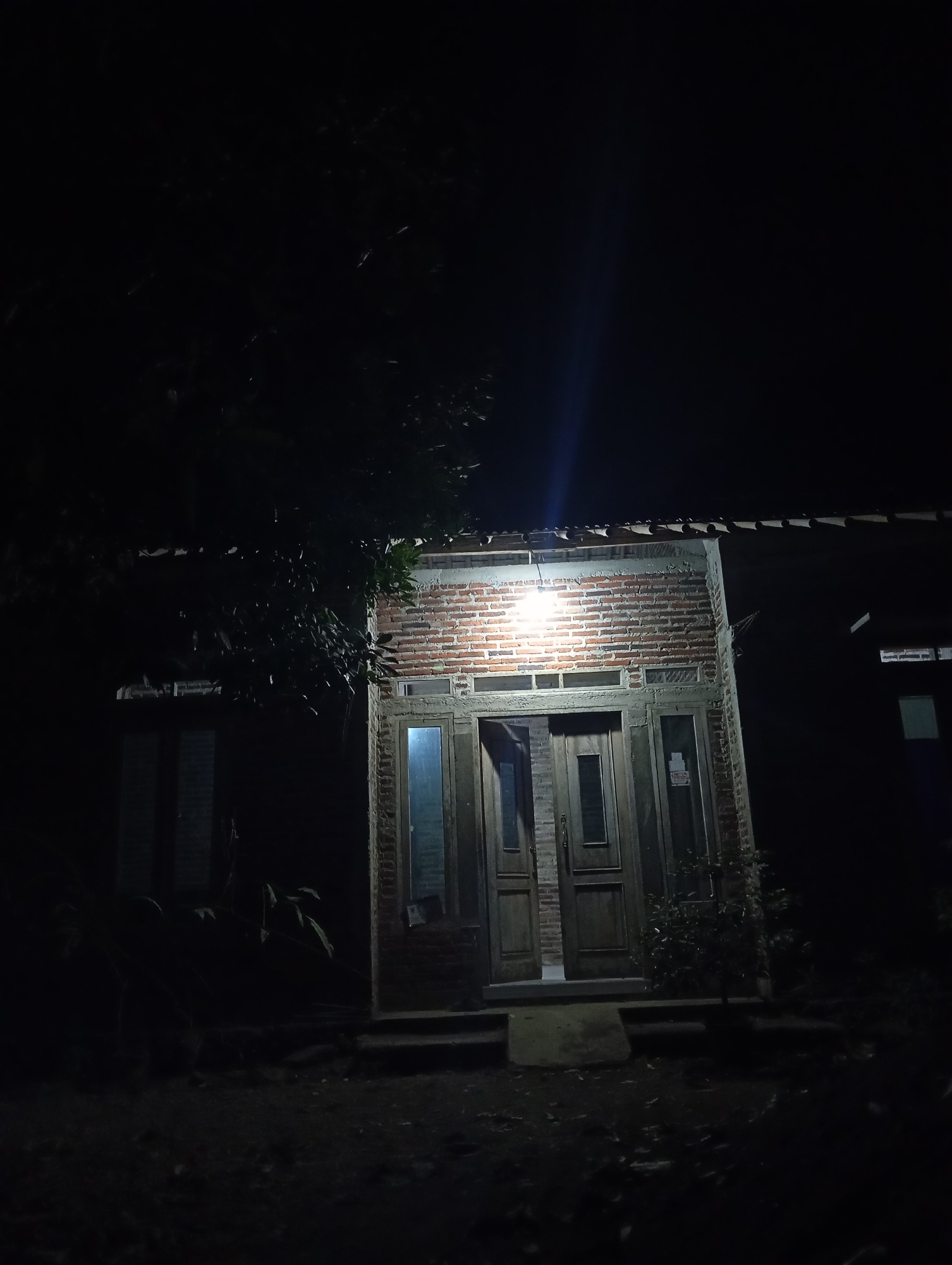 The image depicts the front entrance of a house at night. The house has a brick facade around the entryway, with a wooden double door and two glass sidelights. A light fixture above the door illuminates the area, casting a bright beam upwards. The surrounding environment is dark, with shadows of trees visible on the left side of the house. The ground in front of the house appears to be dirt or gravel. The scene is a residential exterior in Kedungbenda, Indonesia.