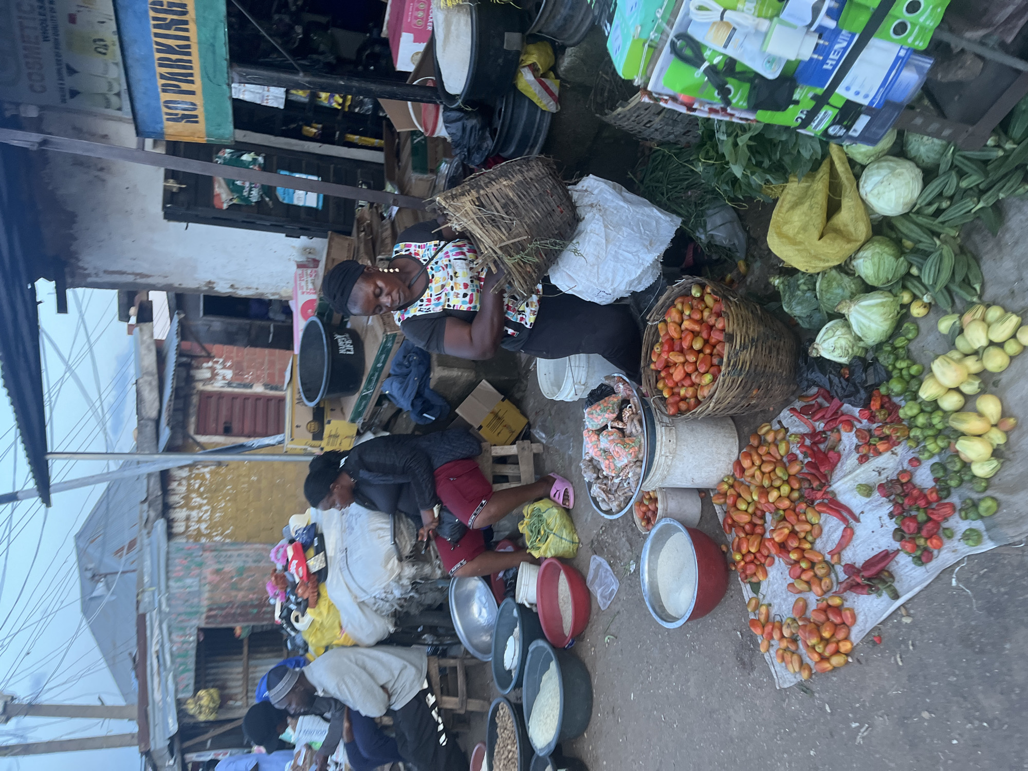 The image depicts a bustling market scene in the city of Gura Top, Nigeria. The foreground is filled with an assortment of fresh produce, including piles of tomatoes, red chilies, and green peppers, laid out on a cloth. Various vegetables such as cabbage and okra are also visible. Several large metal bowls contain grains like rice and possibly peanuts.
In the mid-ground, a woman wearing a headscarf and an apron is seated, appearing to sort through a woven basket. Another woman sits nearby, engaged with her phone. Behind them, several individuals, including men and women, are also seated or standing, suggesting active commerce.
The background reveals a somewhat dilapidated market structure with corrugated metal roofs and walls, characteristic of many open-air markets in urban Nigeria. Signs are visible, including one that reads "NO PARKING." The overall environment appears to be a typical street market with goods displayed outdoors and a sense of everyday activity. The lighting suggests it may be early morning or late afternoon.
