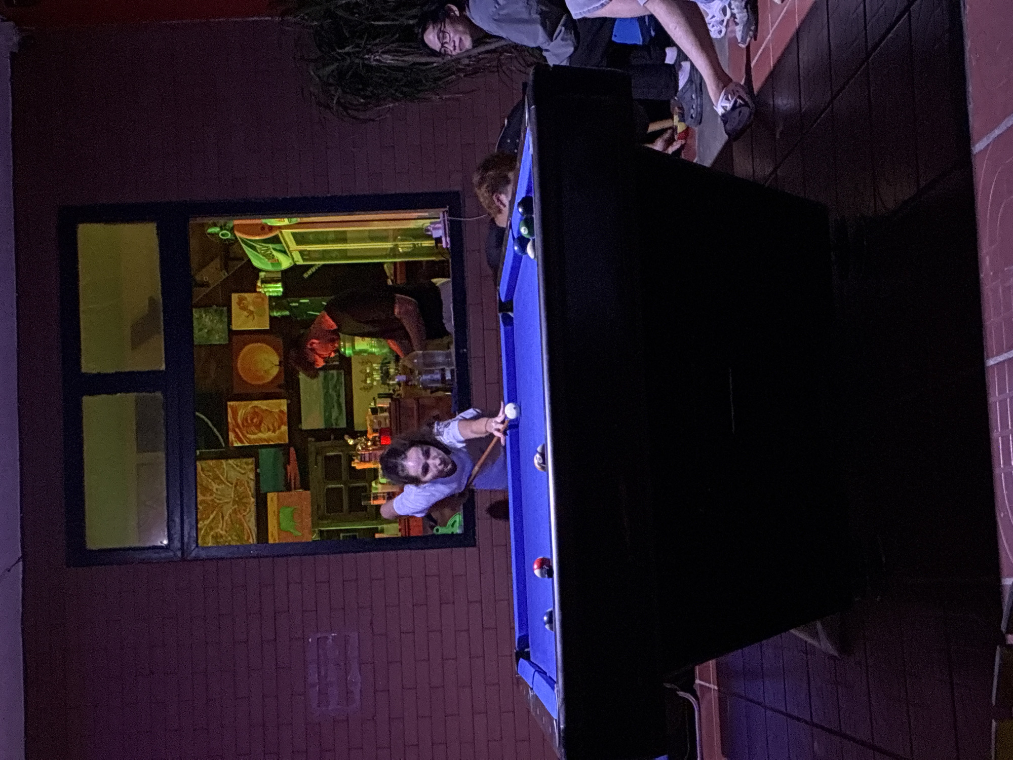 A dimly lit room reveals a pool table in the foreground, with a person in a white shirt leaning over it, cue in hand, about to take a shot. In the background, a window reflects the interior of a bar, where another person can be seen tending to a counter with bottles displayed behind them. Other people are visible around the pool table. The environment seems to be a bar with a pool table area. No specific location in Đà Nẵng, Vietnam, can be identified from the information provided.