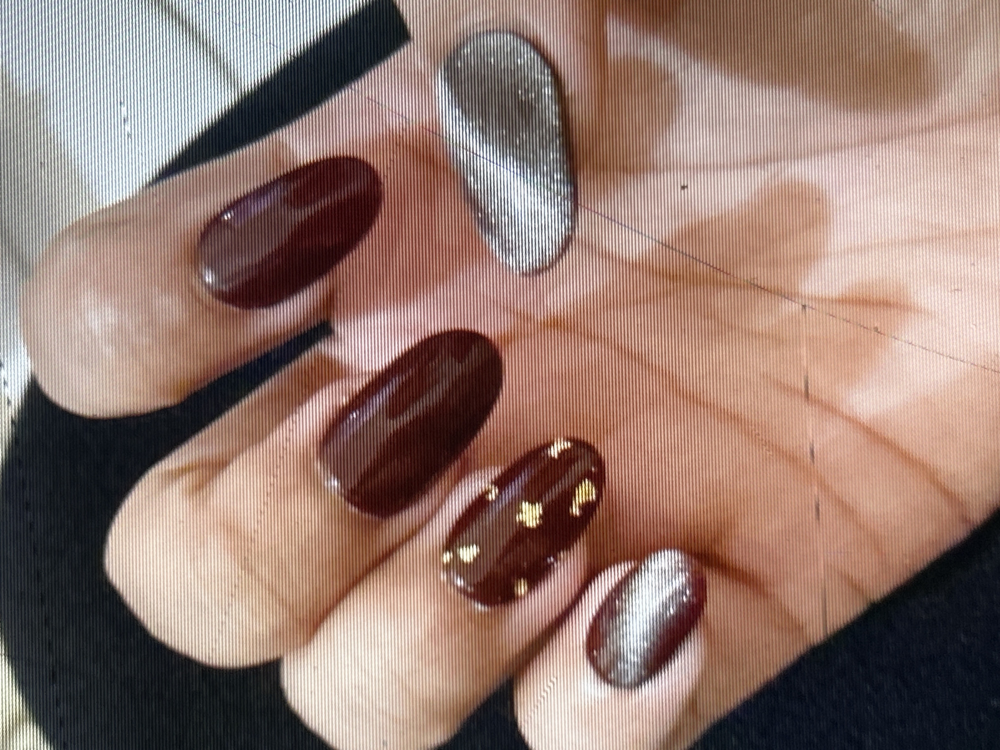 A close-up, high-angle shot shows a person's hand with dark red, oval-shaped fingernails, some with gold star accents, and two nails painted with a silver, holographic polish. The hand is resting on a dark, textured surface, possibly fabric. The lighting is uneven, casting shadows and highlighting the glossy finish of the nail polish. The image quality is degraded by horizontal lines, suggesting interference or a technical issue with the camera or display. There is no discernible information about the location in Depok, Indonesia.