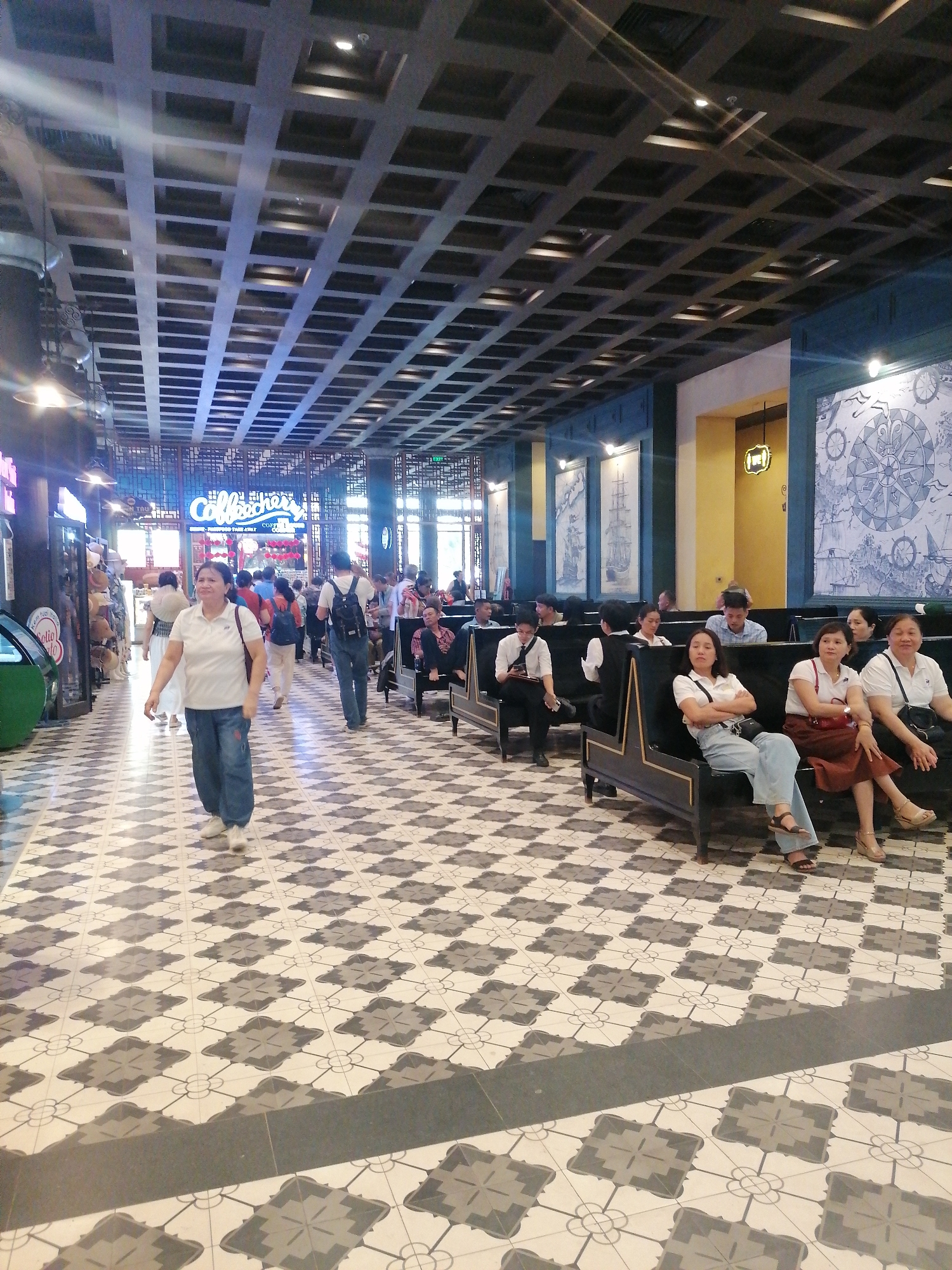 The scene depicts an interior space, likely a shopping mall or a large retail area, in Bai Chay Ward, Vietnam. The flooring is patterned with a repeating geometric design of gray and white tiles. A coffered ceiling extends overhead, creating a grid-like effect.
Several people are present. A woman in a white shirt and blue jeans is walking toward the camera. Other individuals are seen walking in the same direction as well, further into the space. People are seated on benches that line the side of the walkway.
The walls are decorated with paintings and nautical themes, including images of ships and compass roses. Some walls are colored yellow and others are blue. The space appears to be well-lit with artificial lighting. There is also a neon sign for "Coffeocher's". The general atmosphere is busy and public.