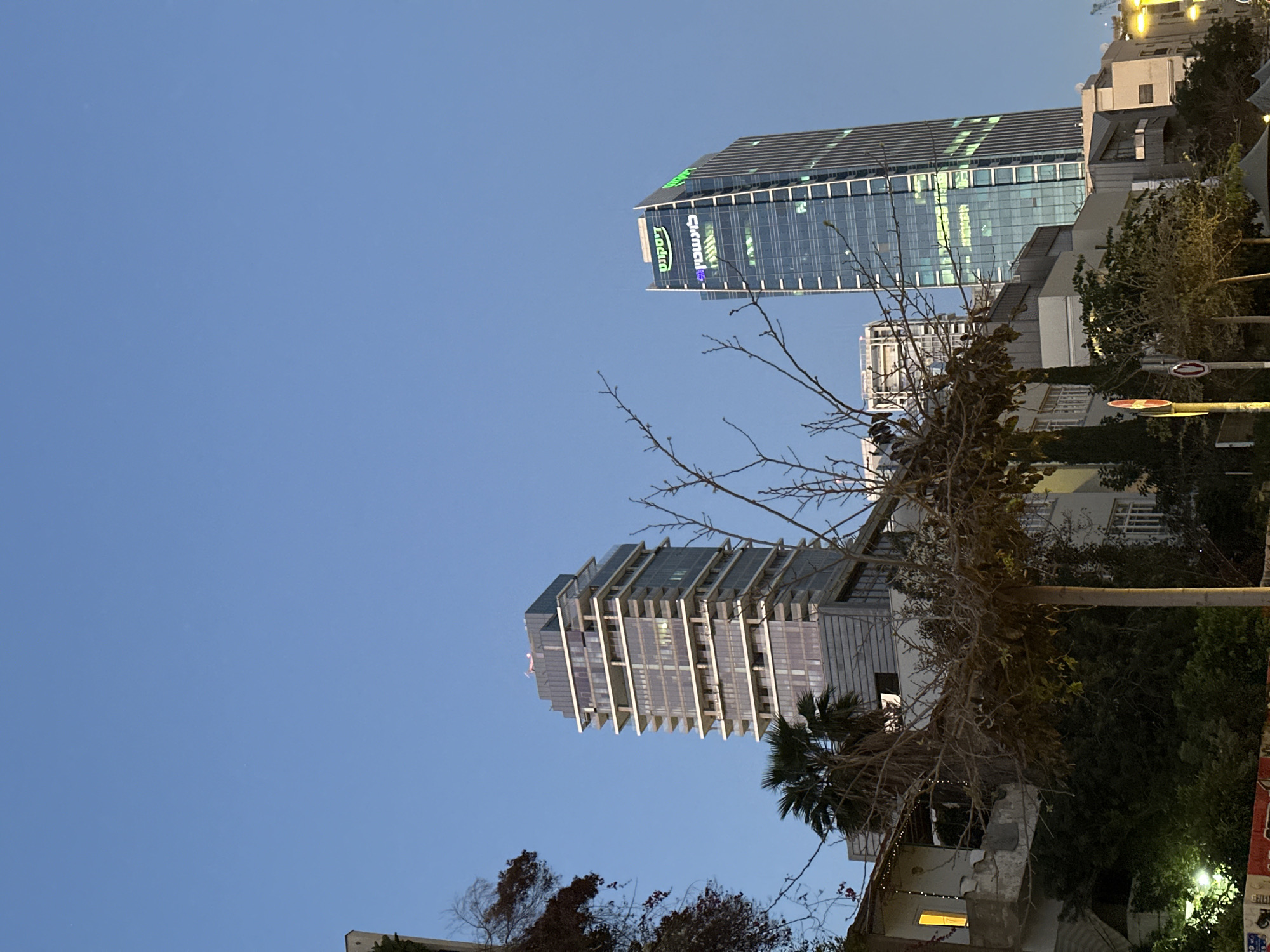 An evening cityscape of Tel-Aviv, Israel, captured with a blue sky overhead transitioning into dusk. The image, rotated counter-clockwise, features two prominent modern high-rise buildings.
In the mid-ground, a tall, sleek corporate building with a glass facade reflects the ambient light. Near its apex, several green illuminated signs are visible. One sign clearly displays "Radix," while another appears to read "Clal," likely referring to Clal Insurance, a prominent Israeli company.
Closer to the viewer and slightly to the left, another modern high-rise stands out with a unique, stepped, and terraced architectural design, featuring balconies or sunshades.
The foreground and lower right section of the image contain darker elements, including dense vegetation with both bare branches and green foliage, suggestive of trees and bushes. A palm tree is also visible. Portions of lower residential or commercial buildings, streetlights, and what appear to be road signs (one red circular sign with a white bar) are subtly illuminated by artificial lights, reinforcing the impression of early evening. The sky is clear, with no clouds visible.
