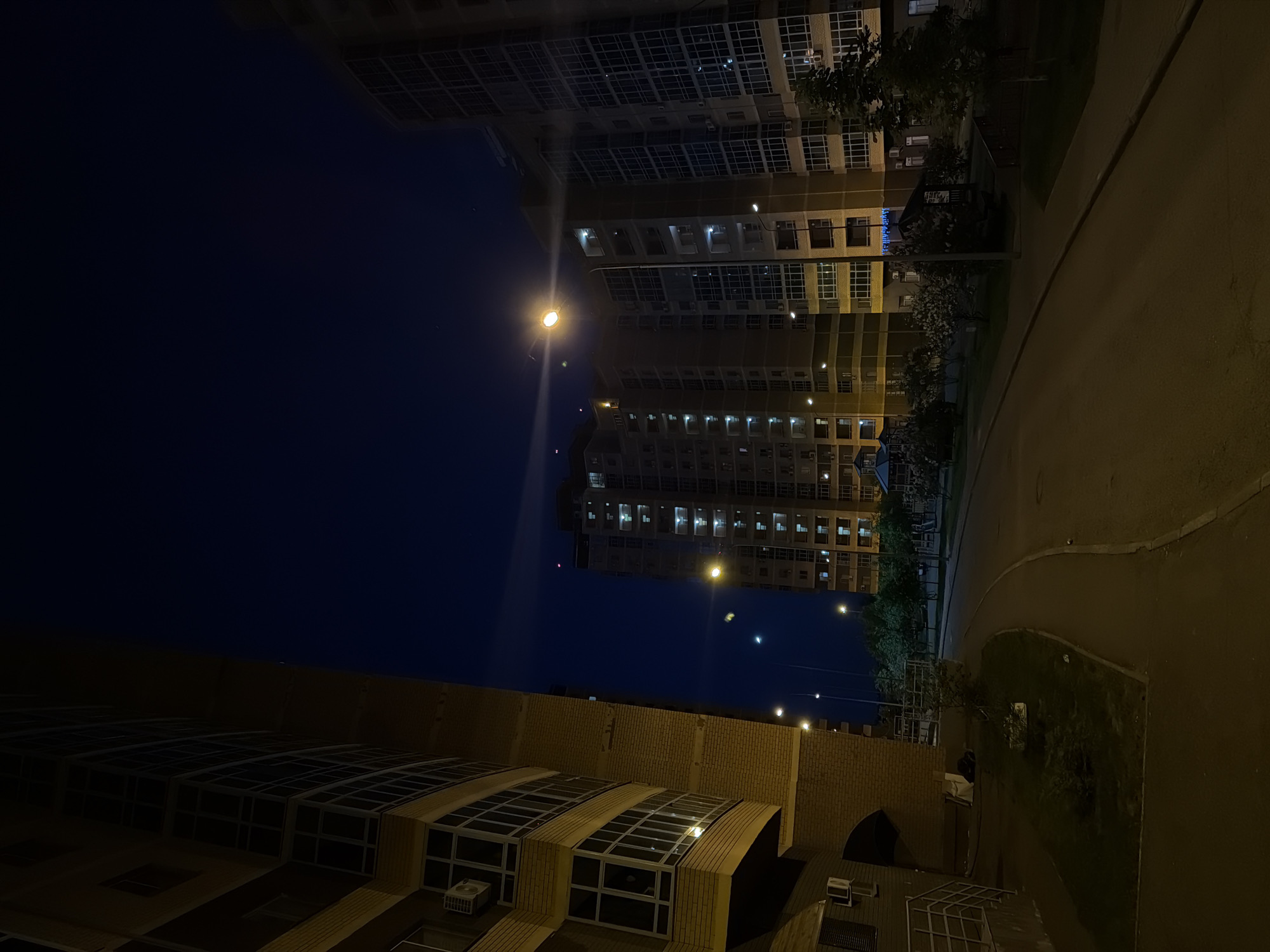 The image shows a nighttime urban scene in Kazan, Russia. In the foreground, there is a low, brick building with multiple windows. Above it, a high-rise apartment building is visible with many illuminated windows. Streetlights are present along the street beside the building. There is a sidewalk adjacent to the building and the road, and some trees are also visible between the building and the sidewalk. The sky is dark.